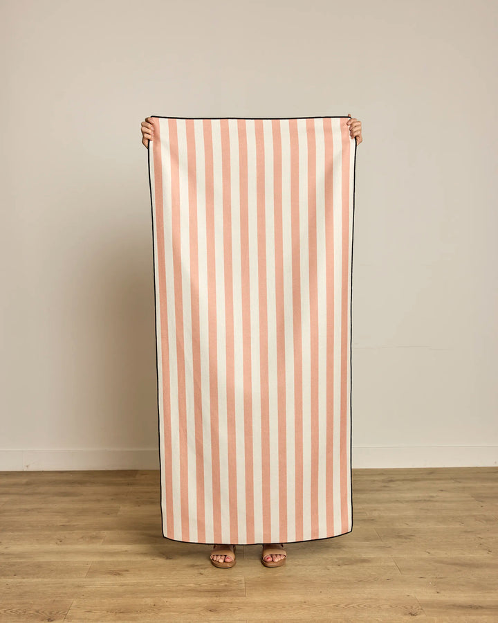 Indoors on a wooden floor, someone holds up Geometry’s Cabana Micro-loop Beach Towel, featuring vertical pink and white stripes. Made from recycled materials, this quick-drying towel covers their body except for their feet and hands.
