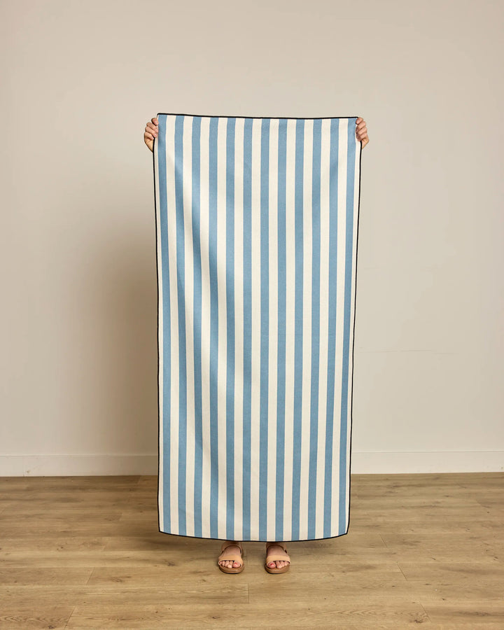 A person indoors on a wooden floor holds up a large rectangular Geometry Cabana towel with vertical blue and white stripes, made from recycled materials, covering most of their body except for their feet.
