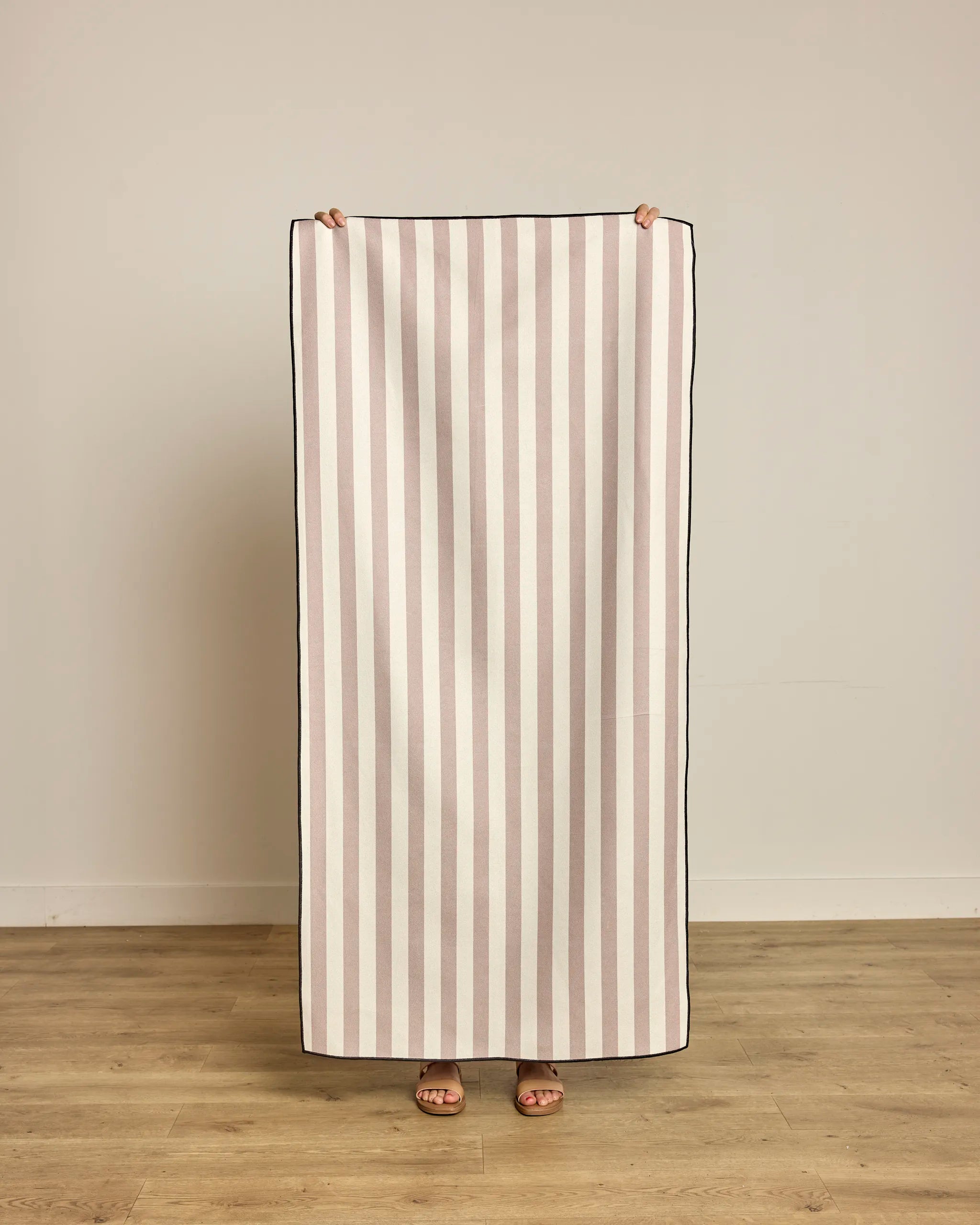 Indoors on a wooden floor, a person holds up the Geometry Cabana Micro-loop Beach Towel, featuring vertical beige and white stripes. Quick-drying and made from recycled materials, the towel hides all but their hands and feet.