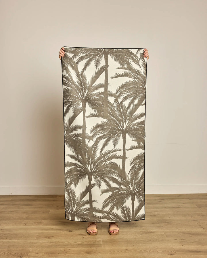 A person stands indoors on a wooden floor, holding up Geometry’s Caligo Brown quick-drying beach towel made from recycled materials, featuring a brown palm tree pattern and concealing their body except for their feet in sandals.