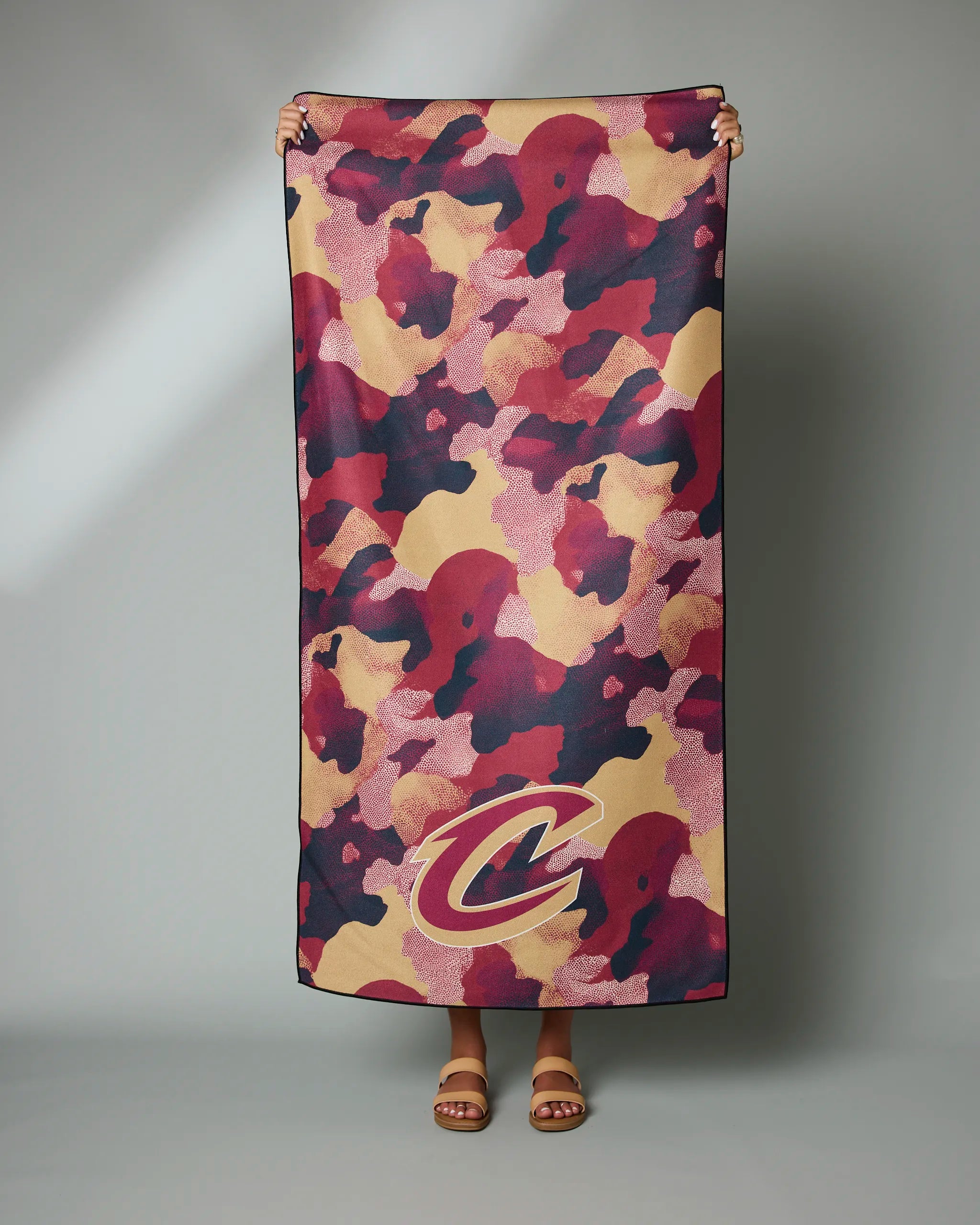 Only the hands and feet of a person are visible as they hold up the Geometry Cavaliers Camo towel, featuring a red, tan, and black camouflage pattern with a large Cleveland Cavaliers C logo near the bottom.