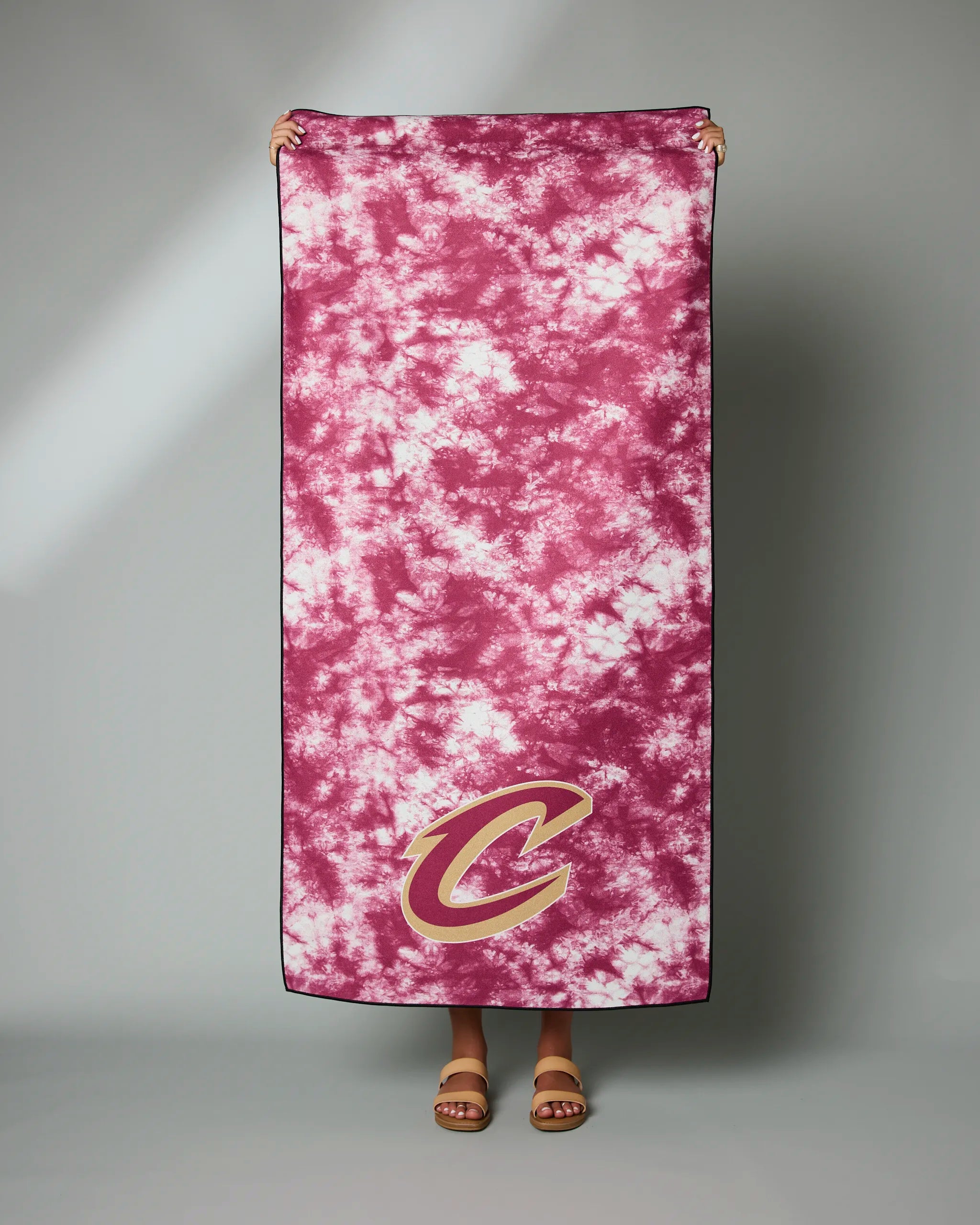 A person holds up the Geometry Cavaliers Flash beach towel, maroon and white tie-dye with a gold and maroon C logo in the corner, against a plain gray background.
