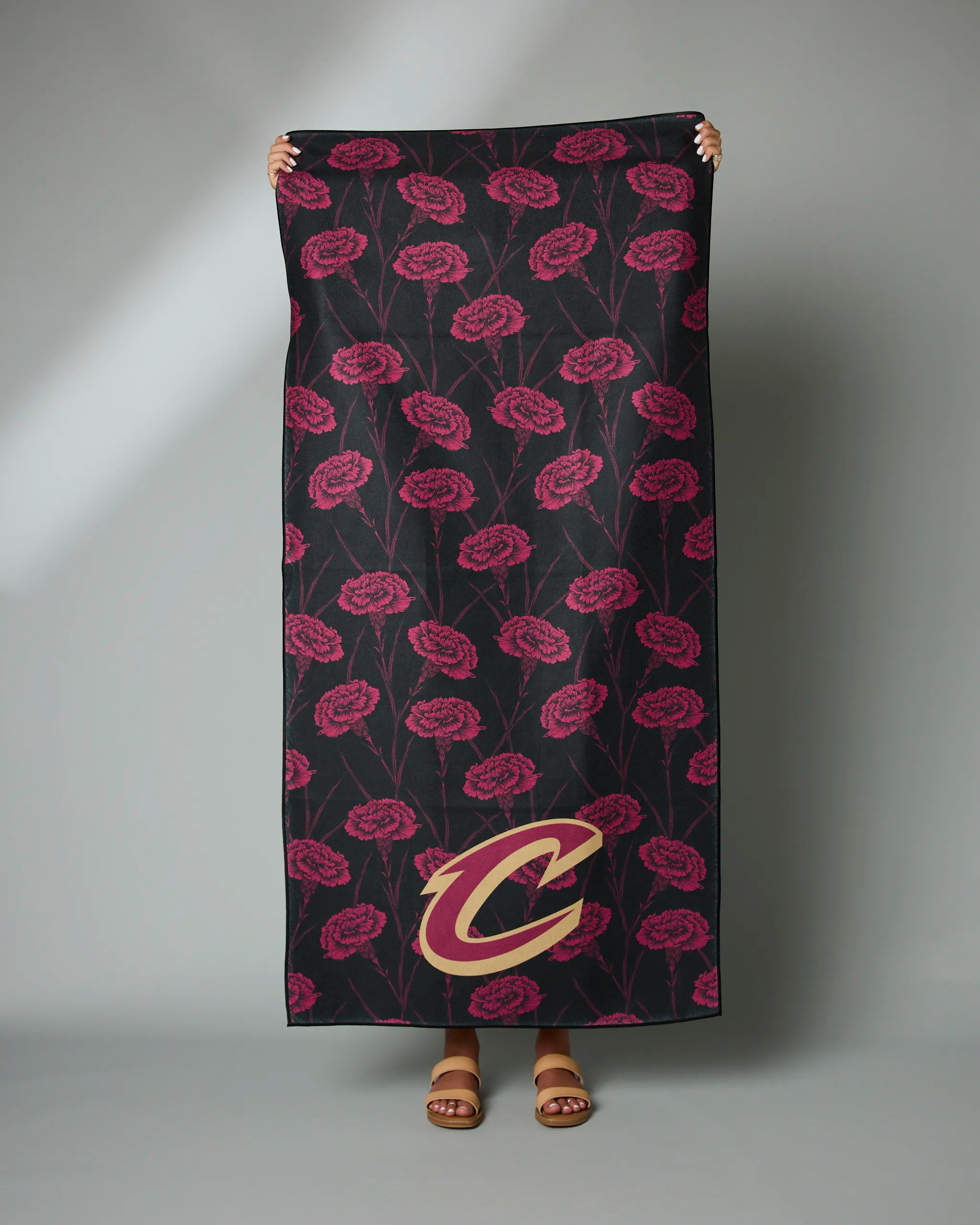 Someone holds up Geometrys Cavaliers Floral towel, featuring red carnations on a dark background with a large gold and red Cleveland Cavaliers C logo in the corner.