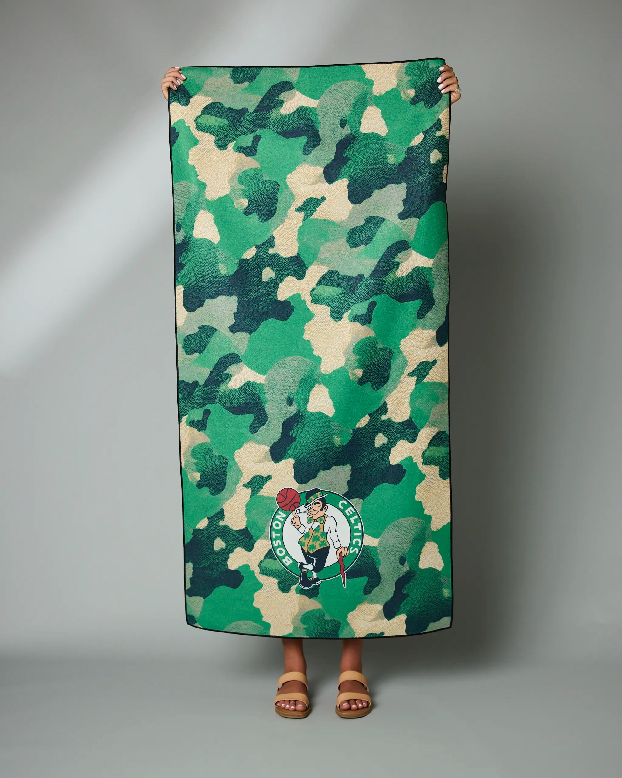 A person wearing sandals holds up the Geometry Celtics Camo towel featuring a green camouflage pattern and Boston Celtics logo, set against a plain gray background.