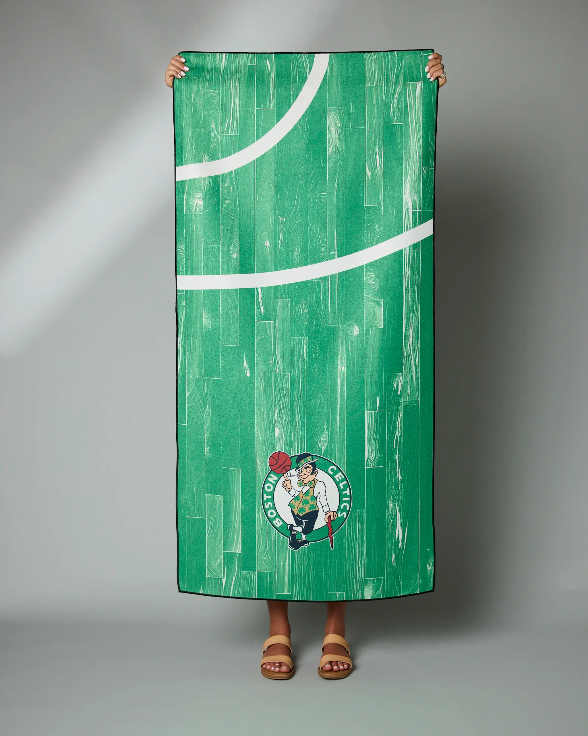 A person holds up the Geometry Celtics Zone towel, styled like a basketball court with the Boston Celtics logo near the bottom. Only their hands and sandal-clad feet are visible.
