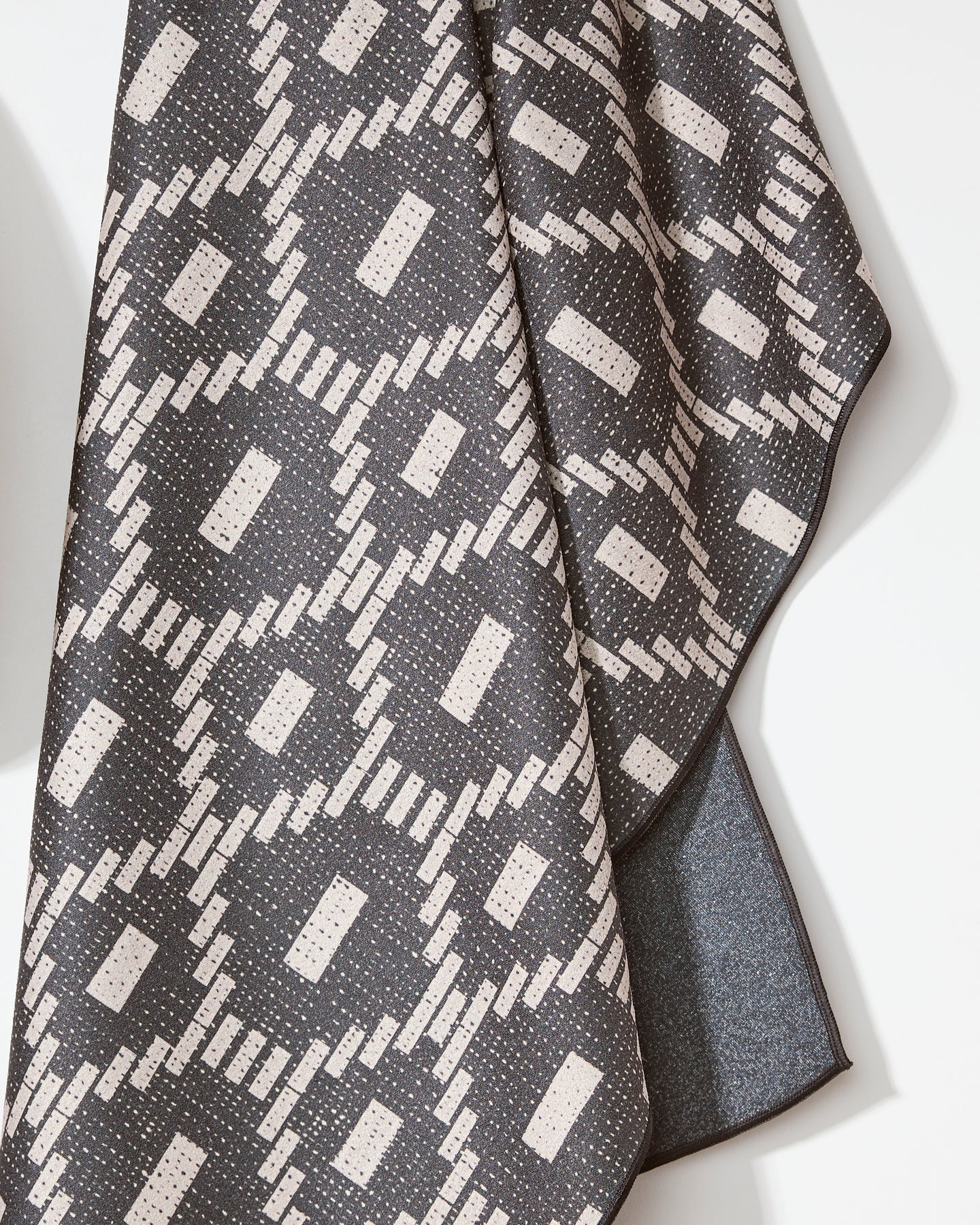 A close-up of the Geometry Charcoal Kiln Micro-loop Beach Towel, featuring a geometric pattern with white rectangles, circles, and diagonal stripes on a dark gray background, made from recycled materials for eco-friendly style.
