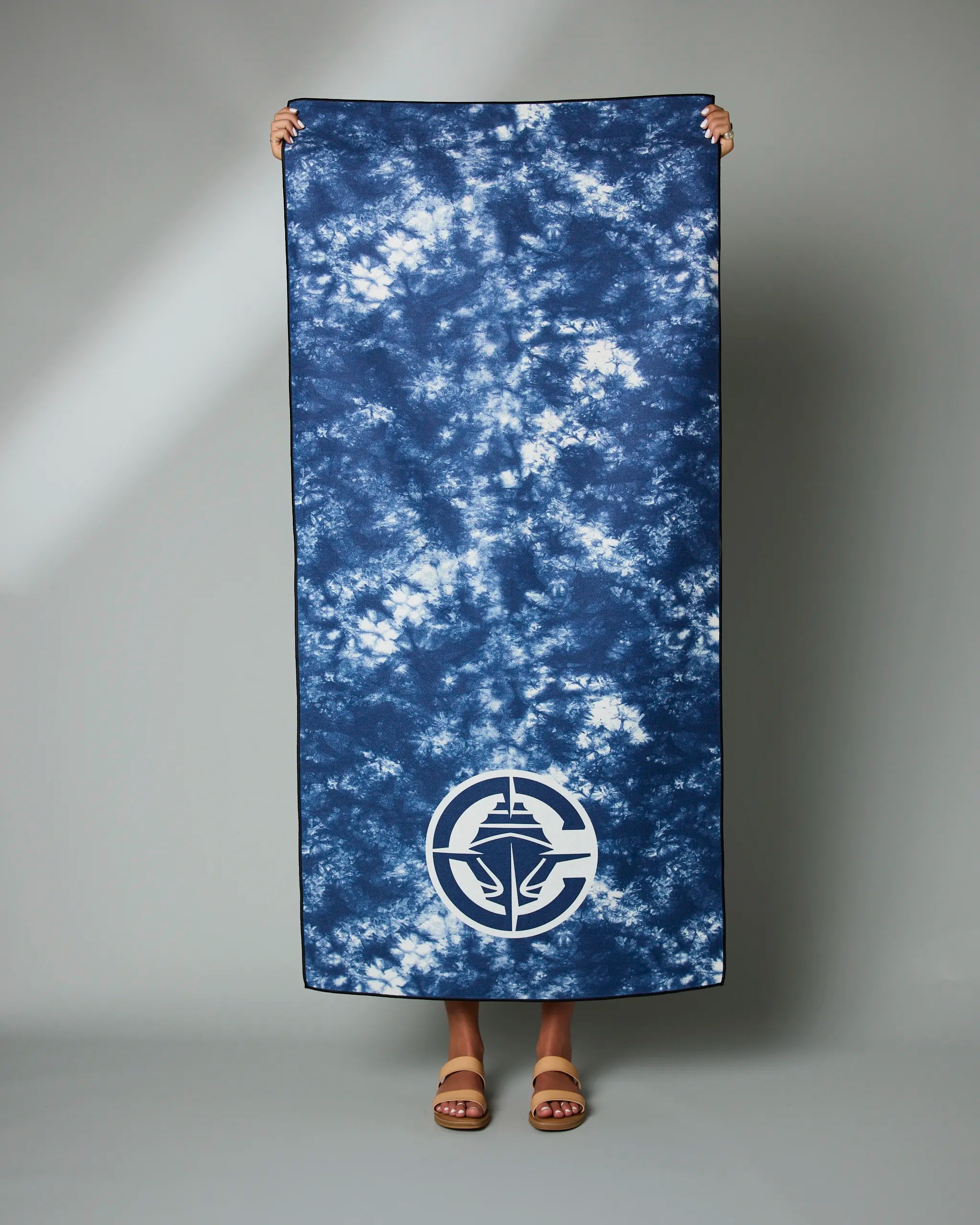 A person holds up the Geometry Clippers Flash large blue and white tie-dye towel with a circular white logo near the bottom, standing against a plain gray background. Only their hands and sandal-clad feet are visible.