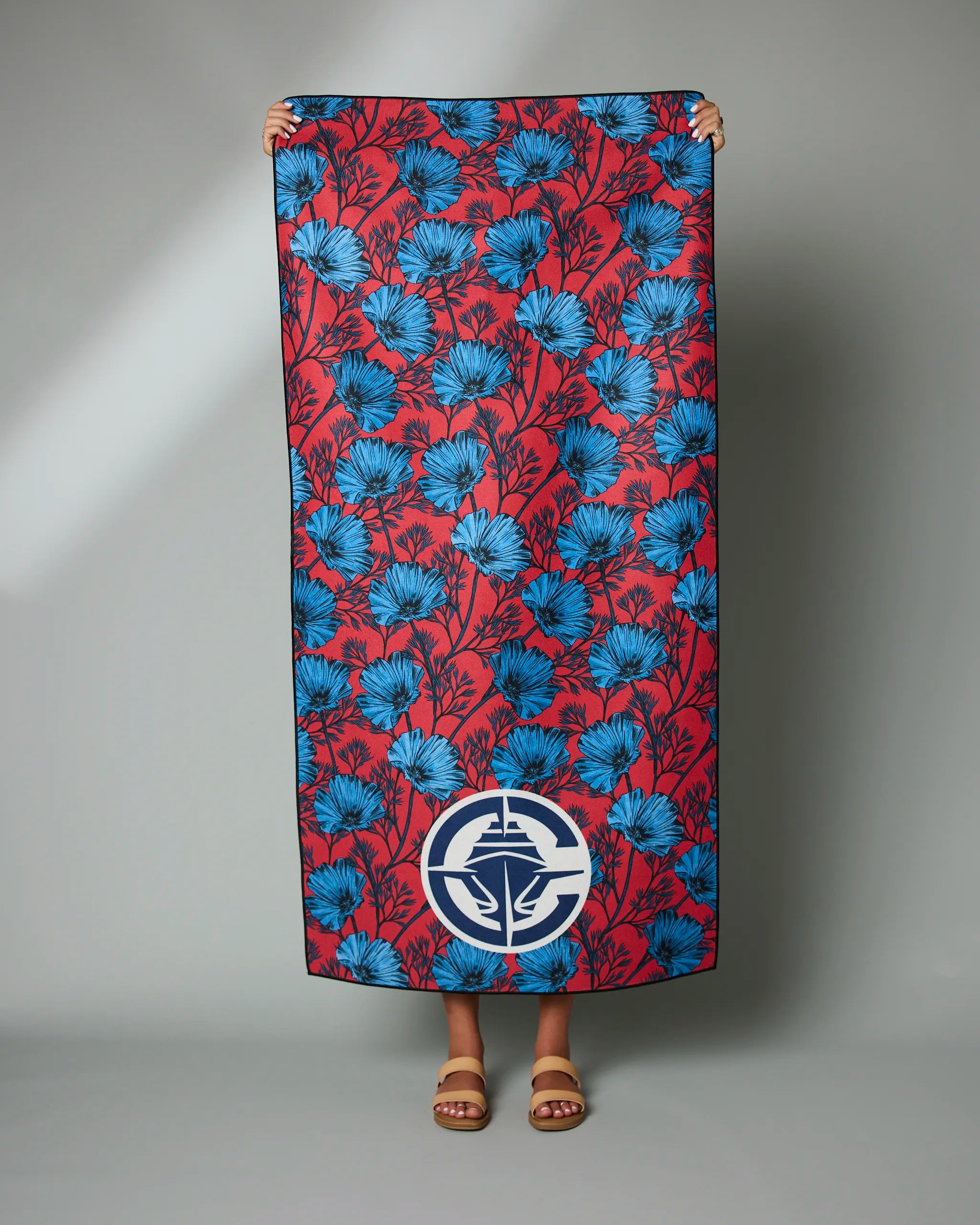 A person holds the Geometry Clippers Floral towel, featuring a large red design with blue floral patterns and a white circular emblem near the bottom. Only their hands, feet, and sandals are visible.