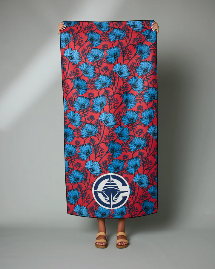 A person holds the Clippers Floral beach towel by NBALAB x Geometry, featuring a blue floral design and white emblem on red. Only their hands and sandal-clad feet show, standing before a plain gray background.
