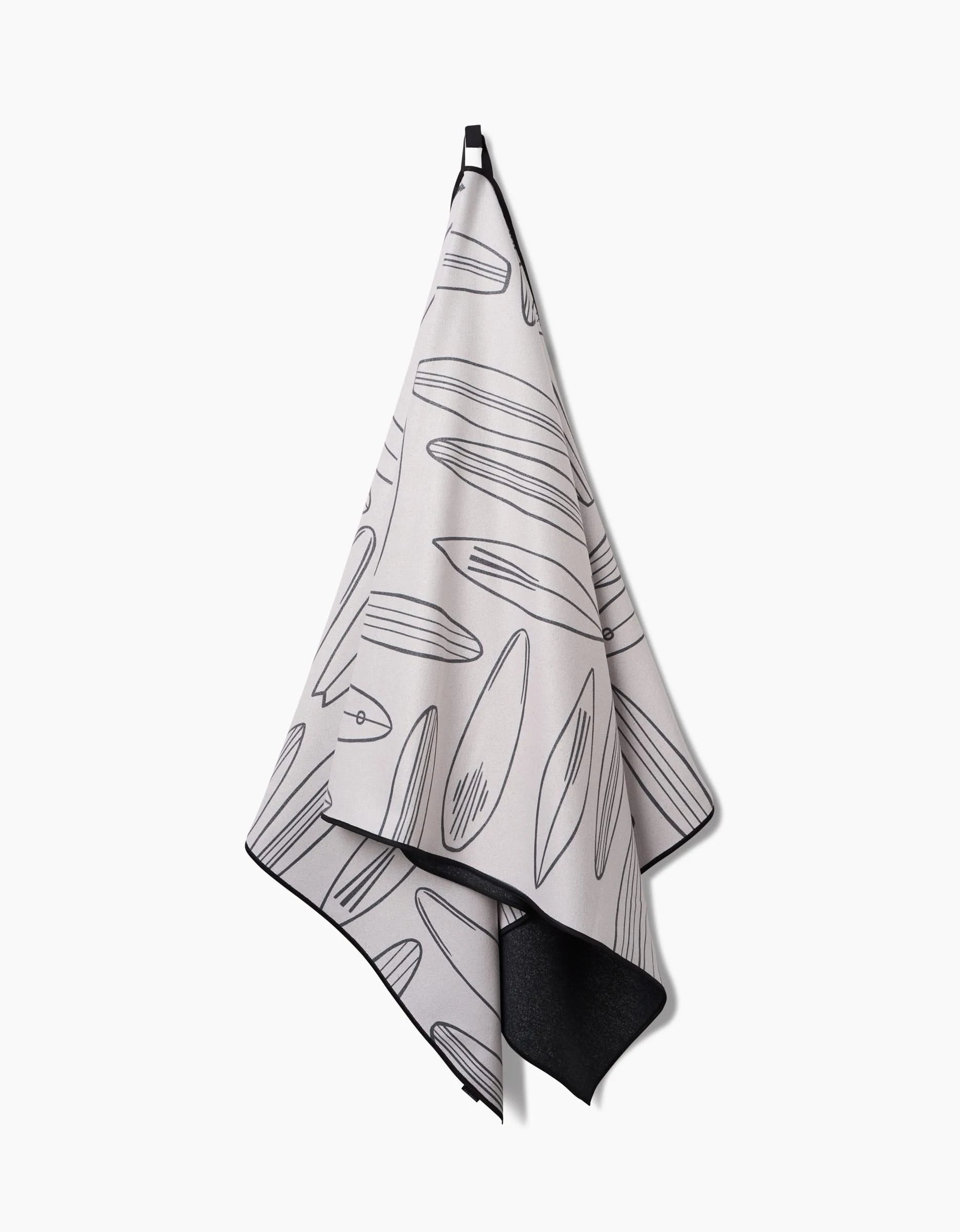 The Hufton Studio Cream Summer Surfing towel, made from quick-drying recycled materials, hangs on a hook with line-drawn surfboards in various shapes. Its light gray color and black edge stand out against the white background.