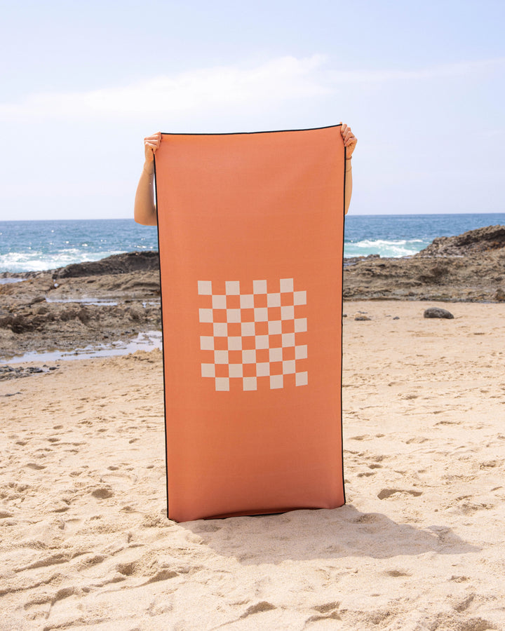 A person stands on a sandy beach, holding up the Geometry Crossplay Peach quick-drying towel with a white checkerboard center. Rocky shoreline and ocean waves can be seen in the background.