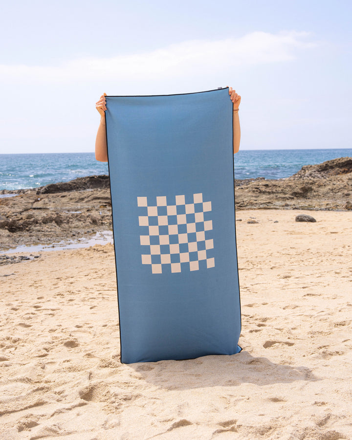 A person stands on a sandy beach holding up a Geometry Crossplay Riviera towel, featuring a white checkerboard design. The ocean and rocky shoreline are visible in the background beneath a clear sky.