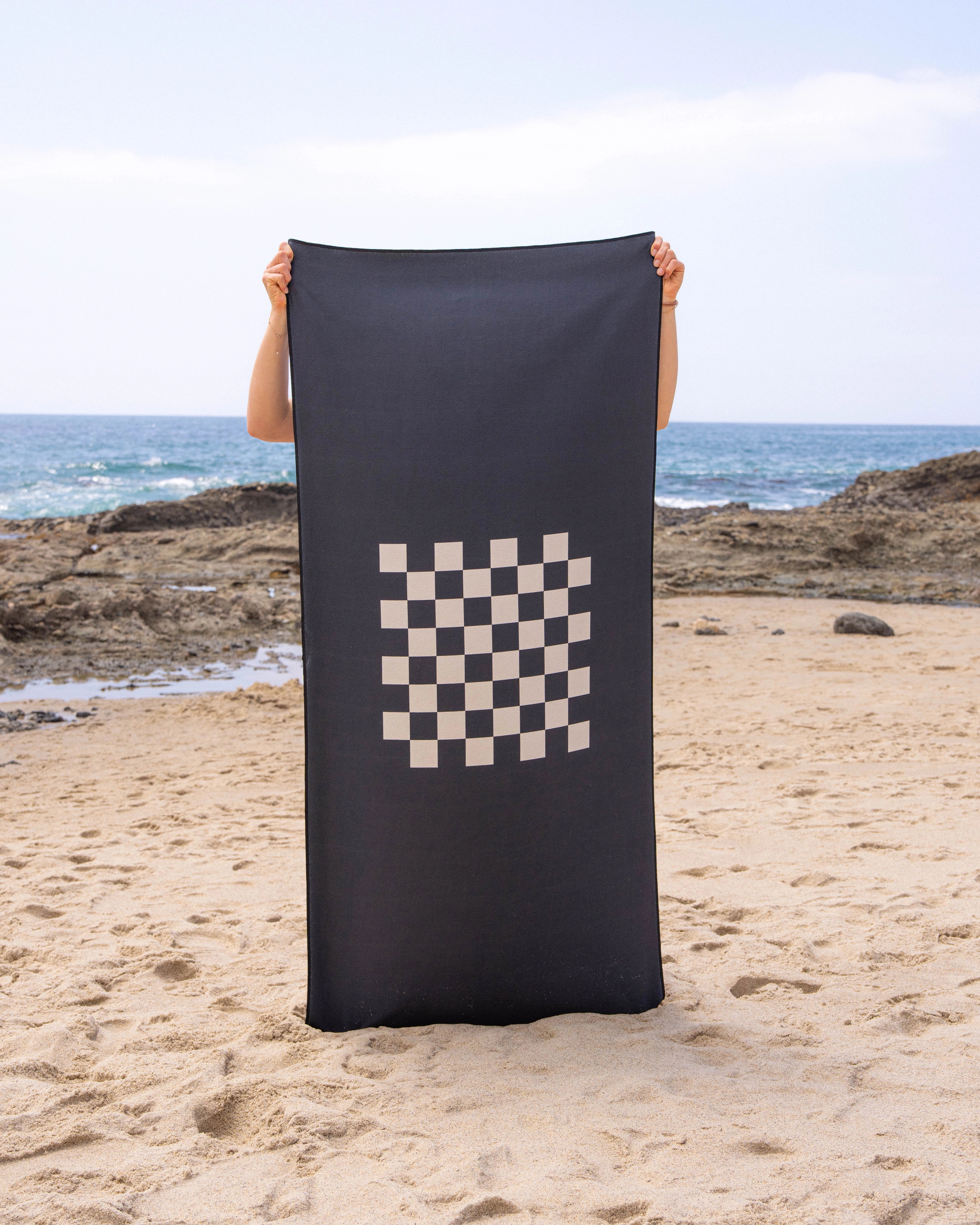 On a sandy beach, someone holds up the Geometry Crossplay Stone quick-drying beach towel featuring a white checkerboard pattern. Rocky shoreline and ocean waves appear in the background beneath a partly cloudy sky.