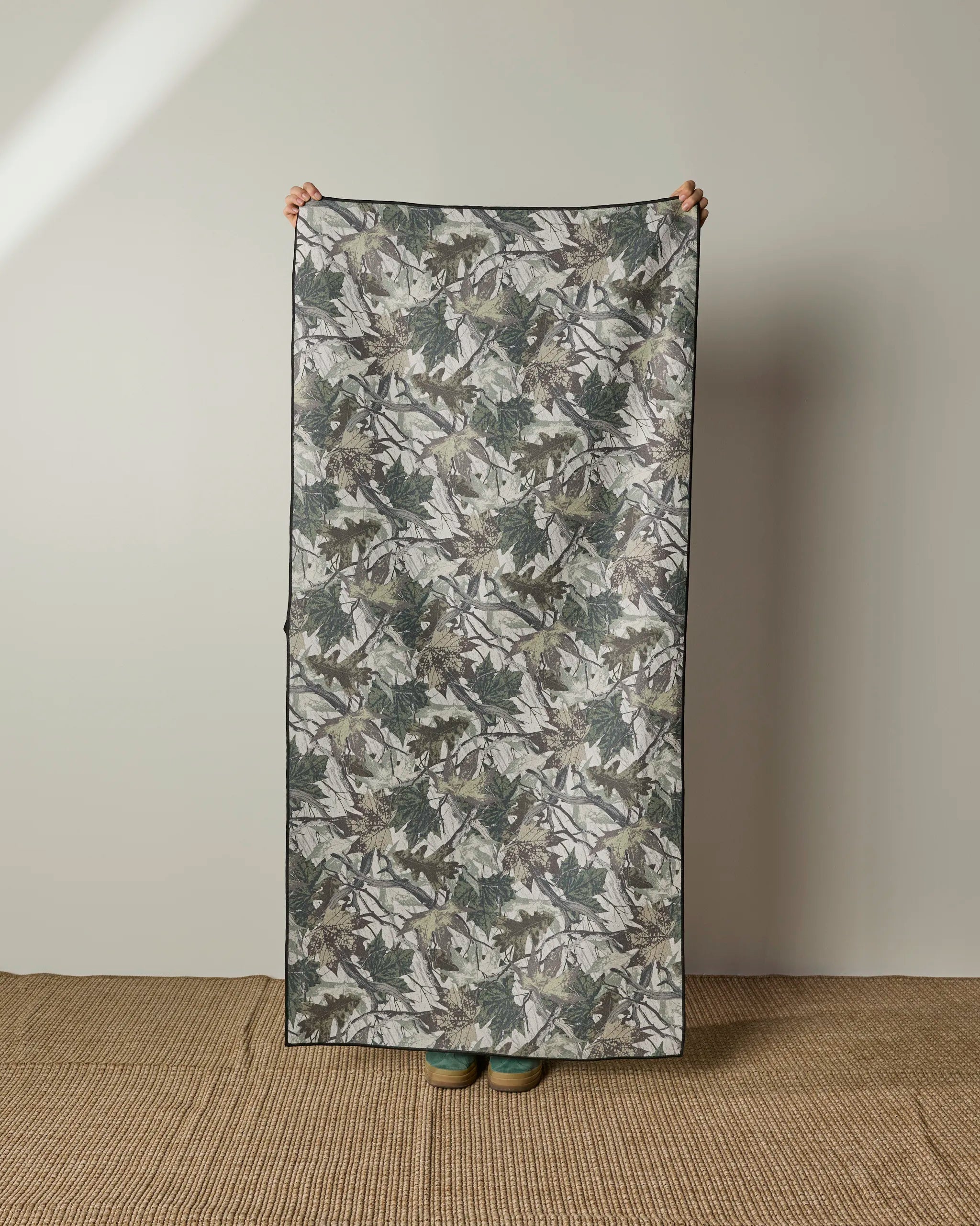 A person stands on a woven rug, holding up the Geometry Dad Camo micro-loop beach towel with a leafy green and gray camo design. Quick-drying and made from recycled materials, this towel covers their body except for hands and feet.