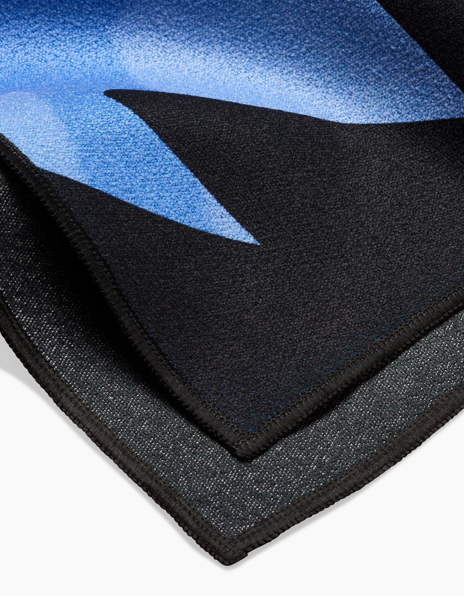 A close-up of Earth by Camila Leão—a folded black towel featuring a blue geometric pattern and stitched edges—highlights the texture and thickness of this quick-drying beach towel.
