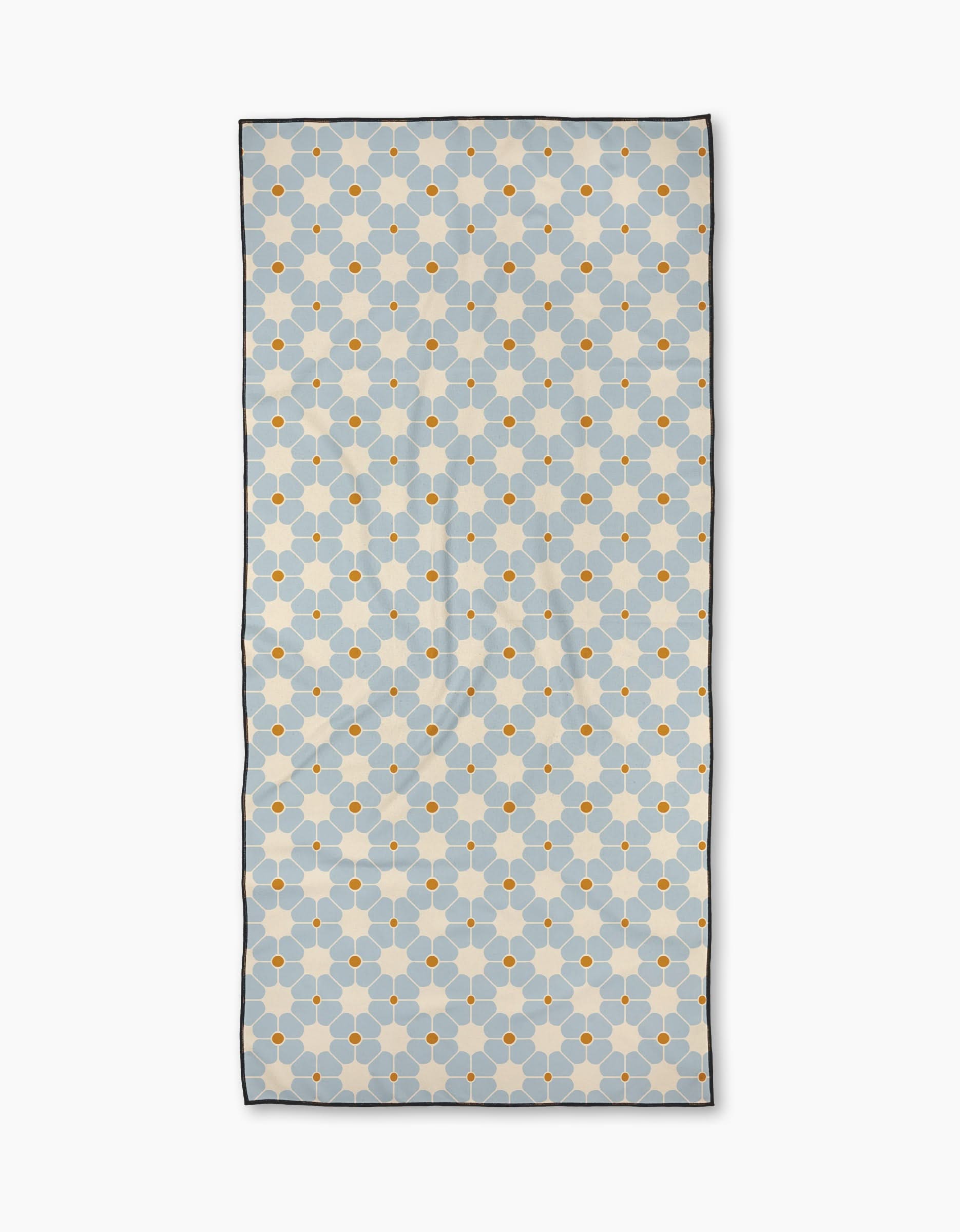 The Flora Tile Light Blue Beach Towel is a rectangular towel with a geometric pattern of light blue shapes and yellow-orange dots on a light background, outlined in dark blue. Made from quick-drying, recycled materials with a thin, dark border.