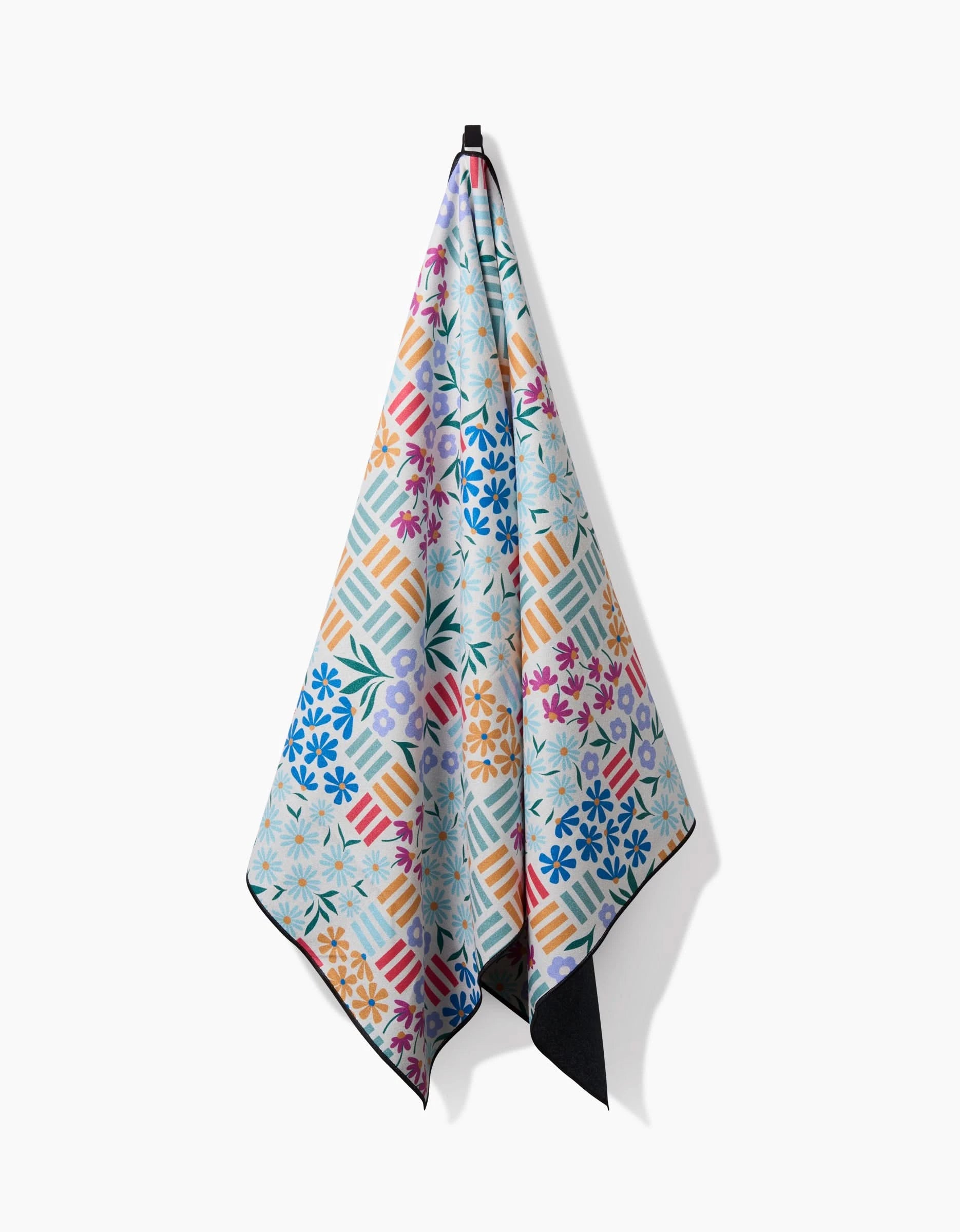 The Floral Garden beach towel by Charlie Rowan Designs, made from recycled materials and featuring a vibrant geometric and floral pattern, hangs against a plain white background.