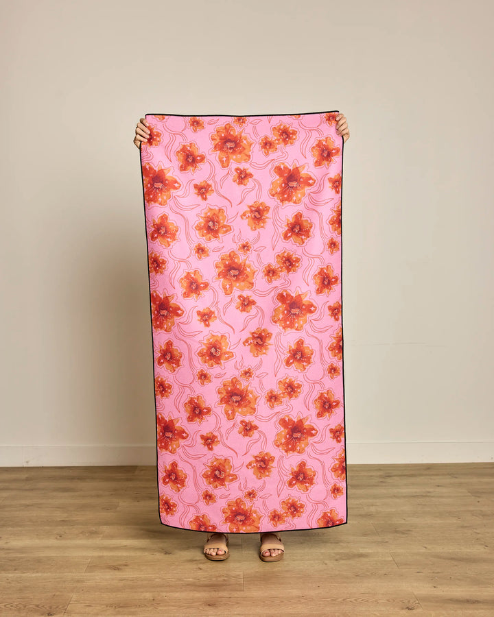 A person stands on a wooden floor holding up a rectangular Geometry “Floral Star Burst” Micro-loop Beach Towel, featuring an orange and red floral pattern. Made from recycled materials, the quick-drying towel obscures their body; background is beige.
