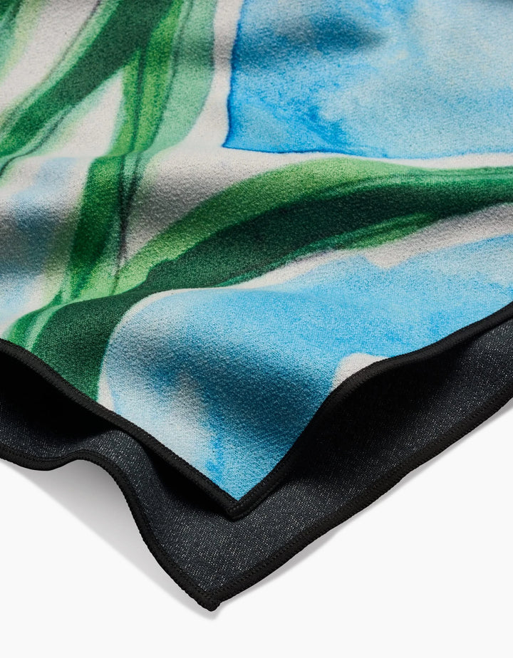 A close-up of the Skye Walker Golden Poppy Flowers beach towel, made from recycled materials, featuring a watercolor-like pattern in blue, green, and white with a black edge, displayed against a white background.