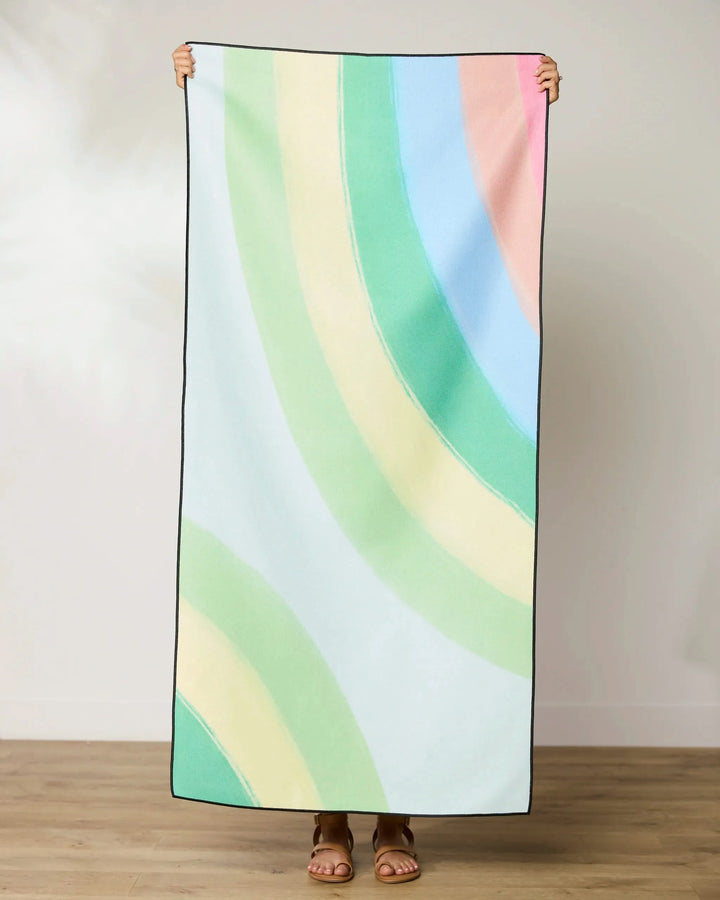 Indoors on a wooden floor, a person holds up the GEOMETRY Green Rainbow beach towel featuring pastel green, yellow, blue, and pink arcs. The quick-drying towel conceals their face—only their feet are visible.