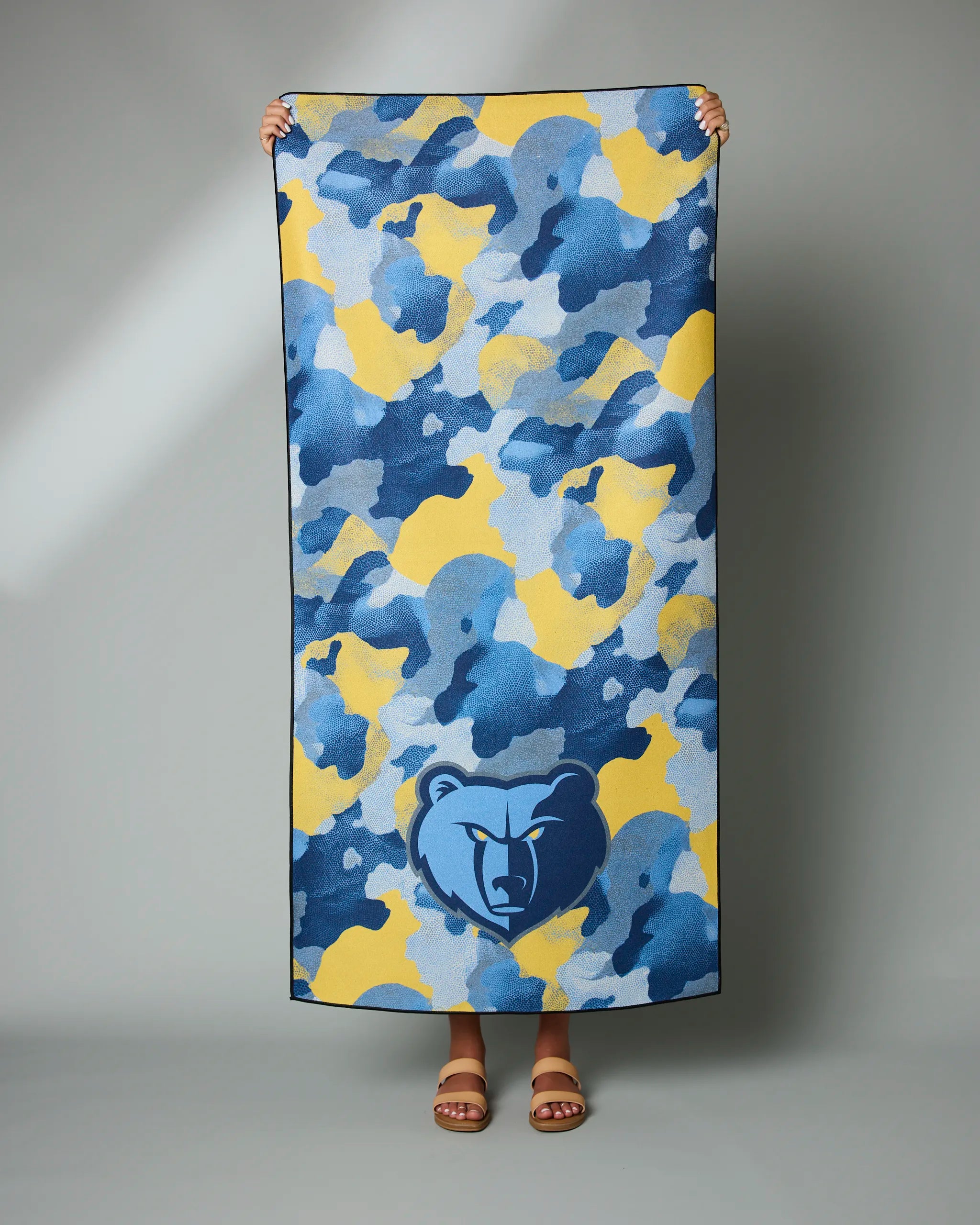 A person holds up the Geometry Grizzlies Camo towel, featuring a blue, yellow, and gray camouflage pattern with the Memphis Grizzlies bear logo at the bottom center. Only their hands and feet are visible.