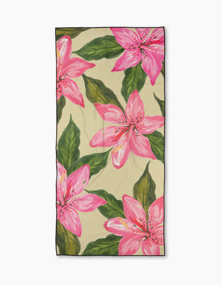 The Geometry Hibiscus Haze beach towel features a beige rectangular design with bold pink lilies and green leaves. Made from quick-drying micro-loop material, it combines comfort, convenience, and vibrant tropical style.