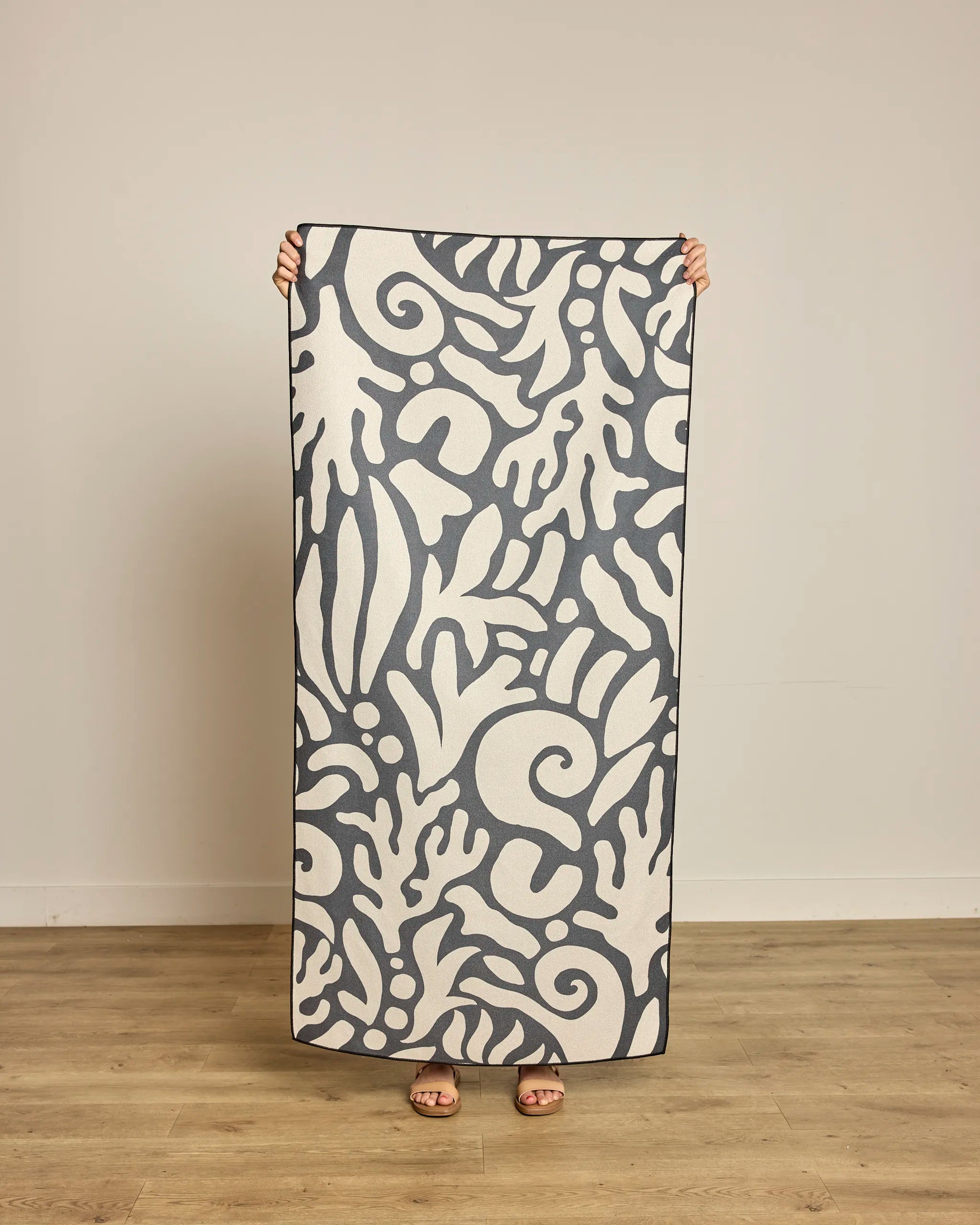 A person stands on a wooden floor, holding up the Geometry Inkwell Reef Beach Towel with a bold black and white abstract pattern that covers their body except for sandal-clad feet. A plain, light-colored wall forms the simple backdrop.