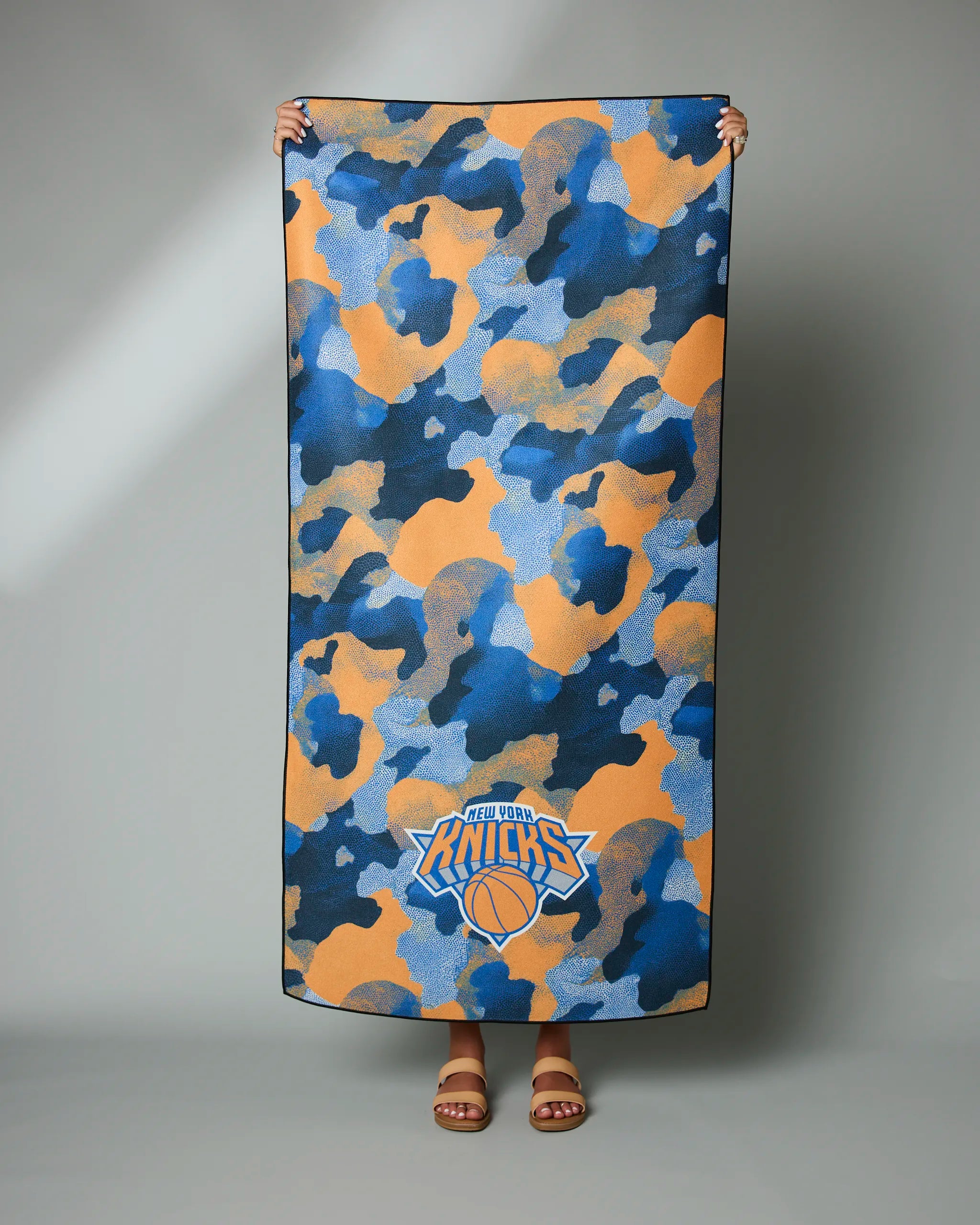 A person holds the Geometry Knicks Camo towel, featuring a blue, orange, and gray camouflage design with the New York Knicks logo at the bottom—only their hands and feet are visible.