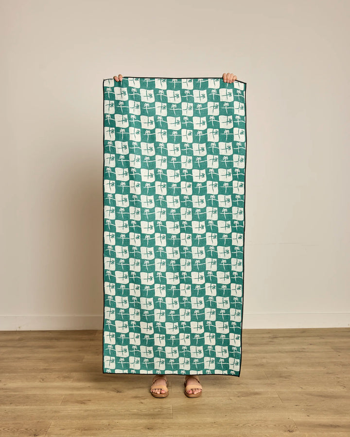 A person stands on a wooden floor, holding up the Geometry La Brisa Green micro-loop beach towel made from recycled materials, covering their body with only hands and feet visible, against a plain light-colored wall.