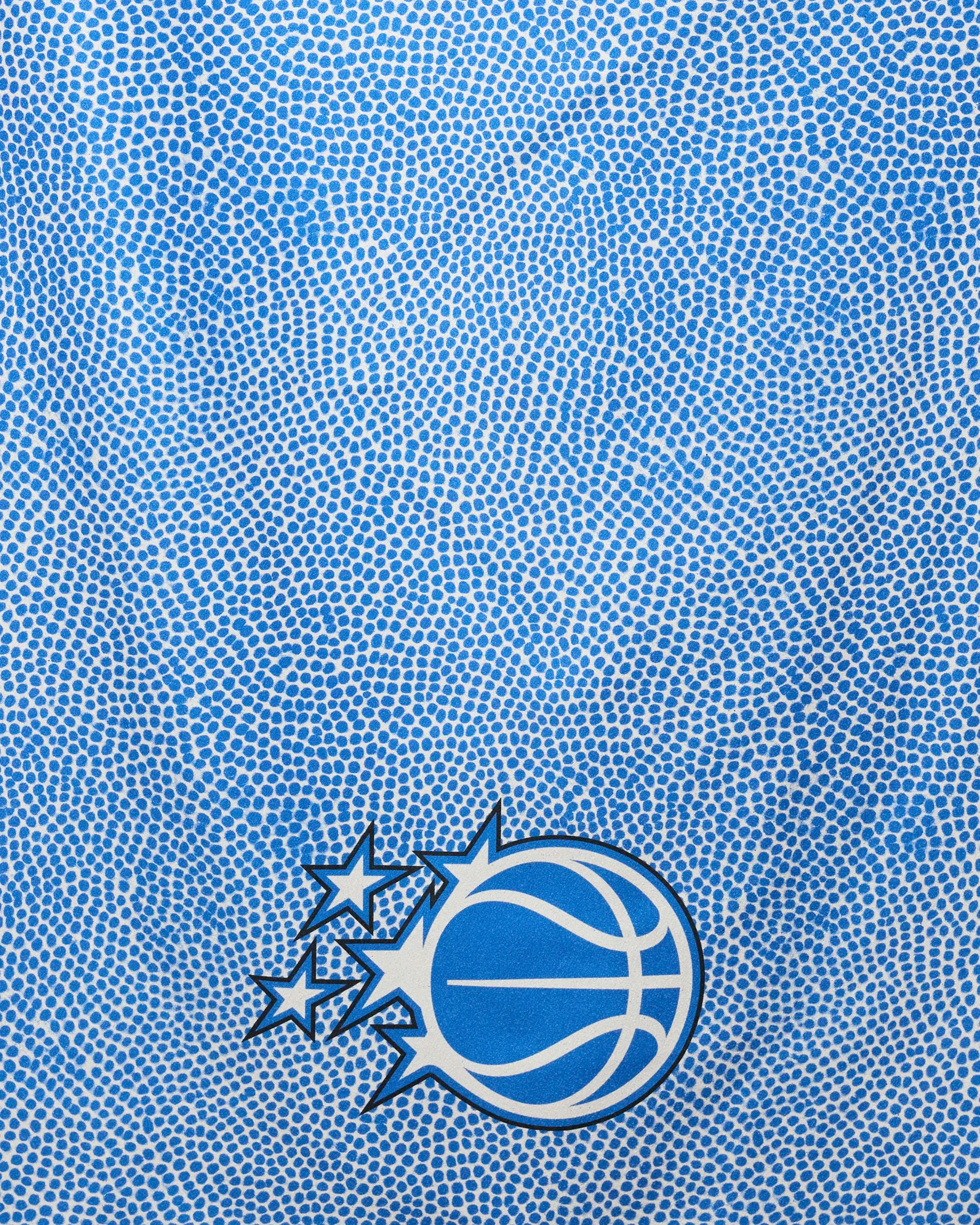 A blue textured background with a basketball pebble pattern. In the bottom left, a stylized basketball with three stars—outlined in white and navy—echoes the NBA energy of the Magic Grip towel by NBALAB x Geometry.