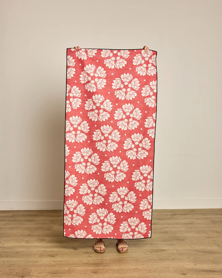 A person stands against a plain wall, holding up the Geometry Maverick Floral pink micro-loop beach towel—featuring a repeating white floral pattern, made from recycled materials, and quick-drying—covering their body except for sandals.