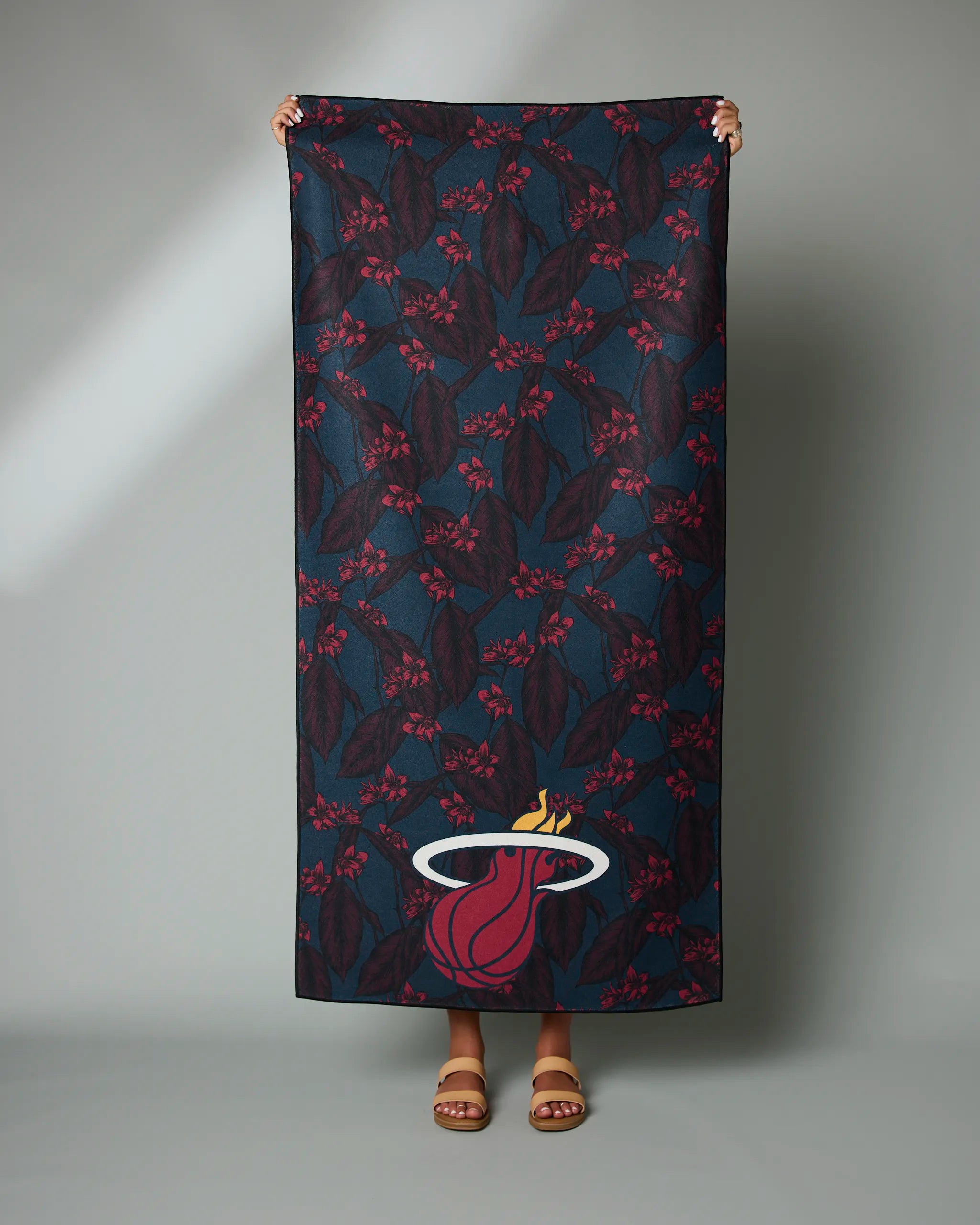 A person displays the Geometry Heat Floral towel, featuring a dark floral design and the Miami Heat logo near the bottom. Only their hands and feet are visible.