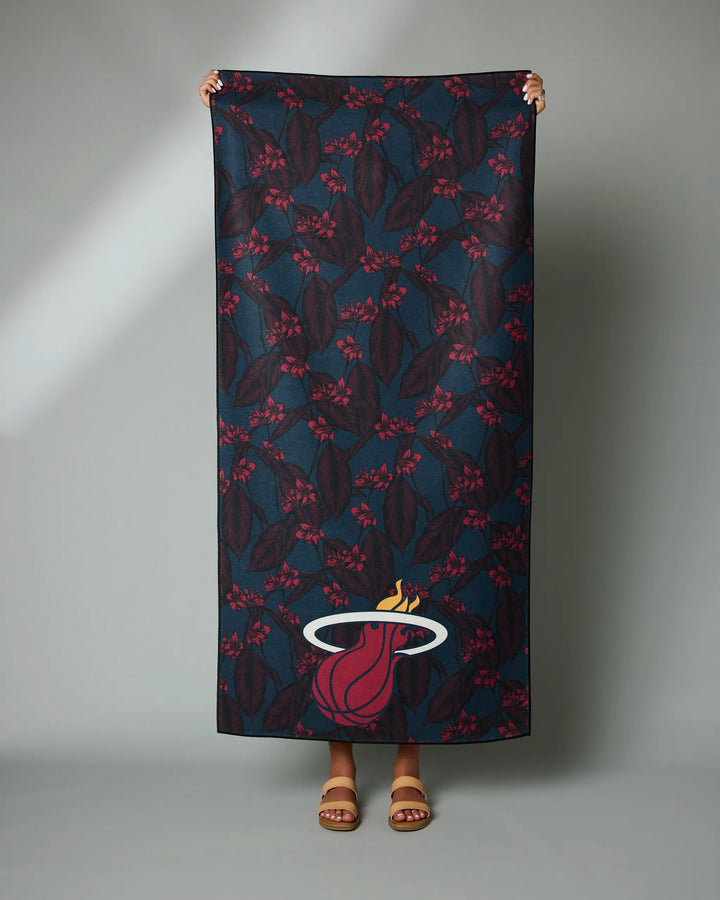 A person holds up the NBALAB x Geometry "Heat Floral" beach towel, featuring a dark floral design and Miami Heat logo, made from recycled materials. Only their hands and feet are visible against a plain gray background.