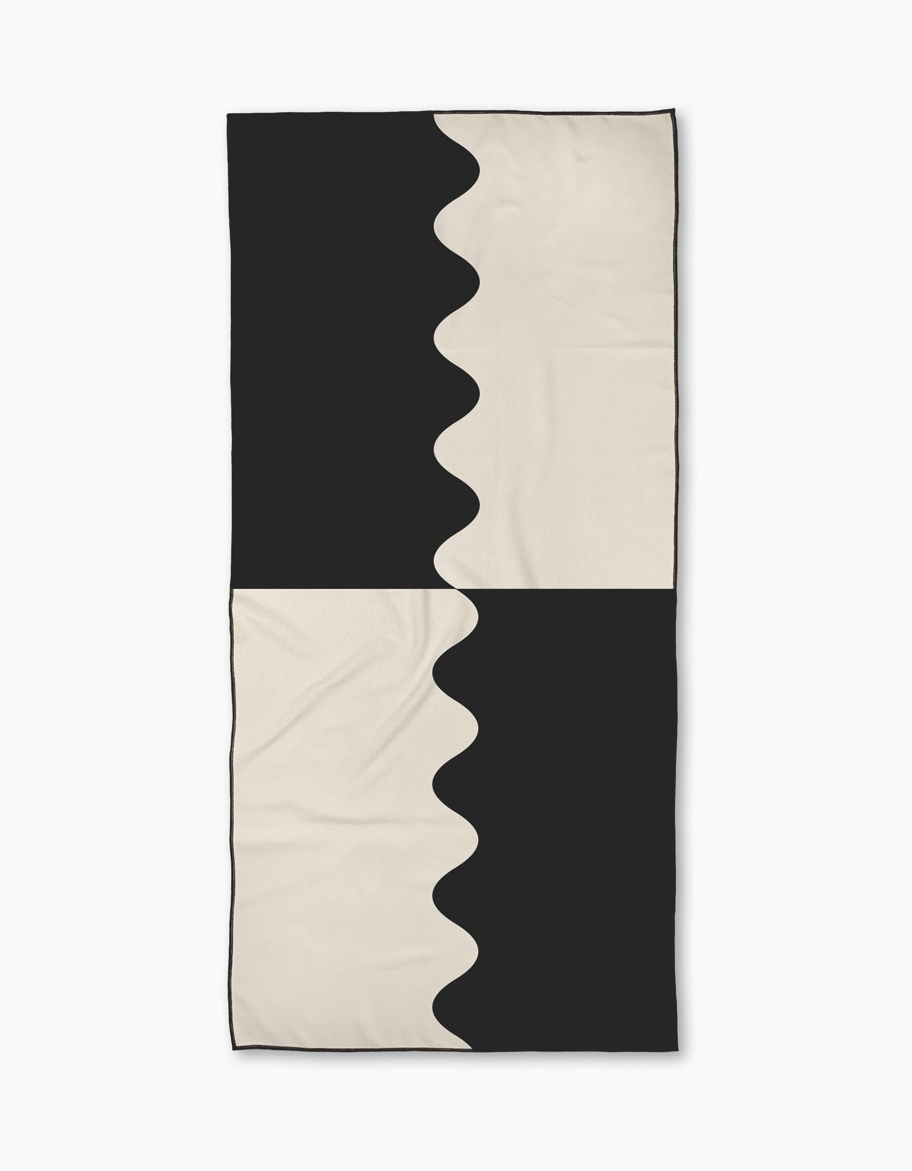 The Geometry Monochrome Waves Micro-loop Beach Towel features a bold black and off-white abstract design with mirrored wavy lines. Made from recycled materials, this quick-drying towel is shown on a white background.