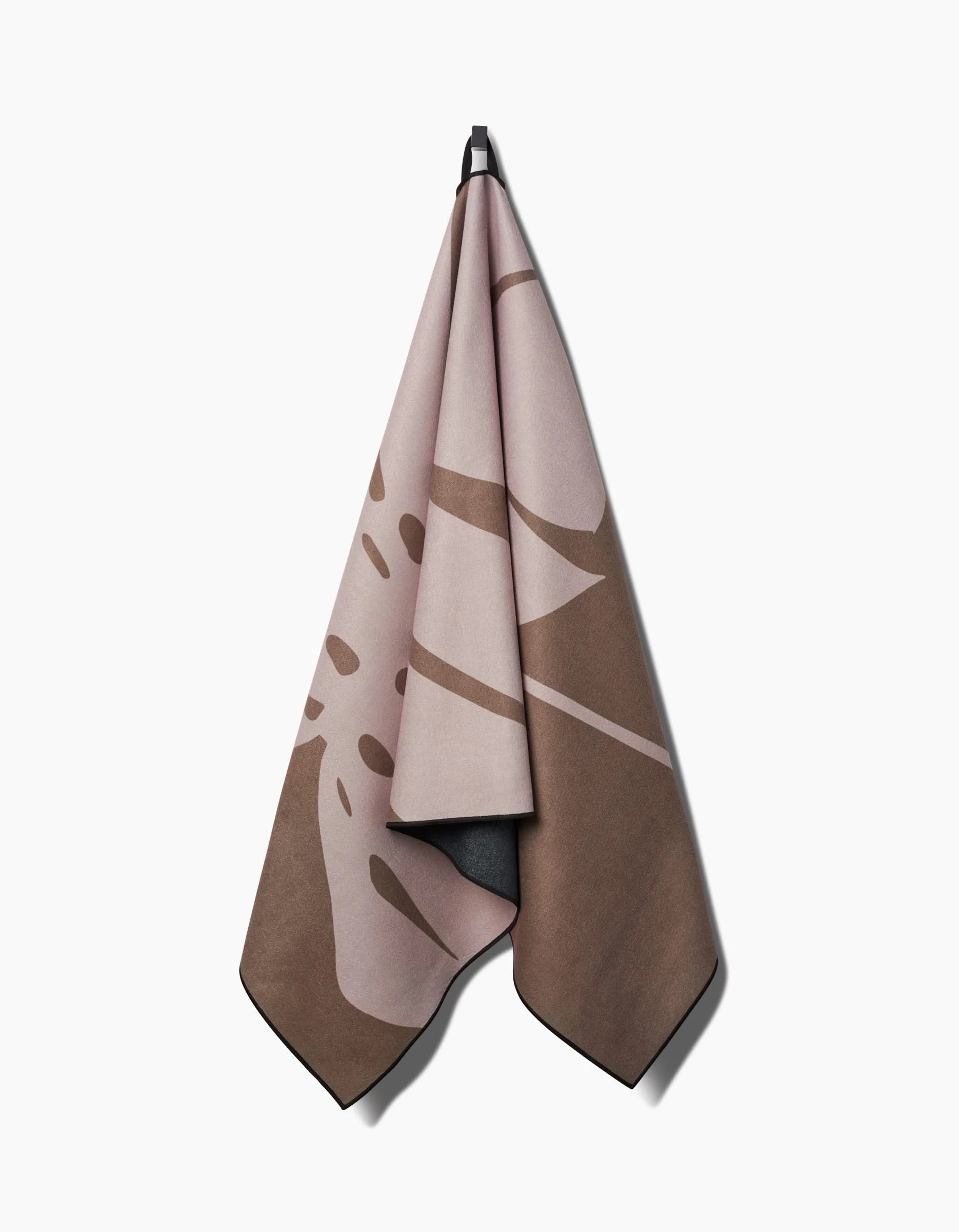 The Monstera Leaf beach towel by PrintedPlant, featuring a brown and beige leafy design and made from recycled materials, hangs neatly from a black hook against a plain white background.