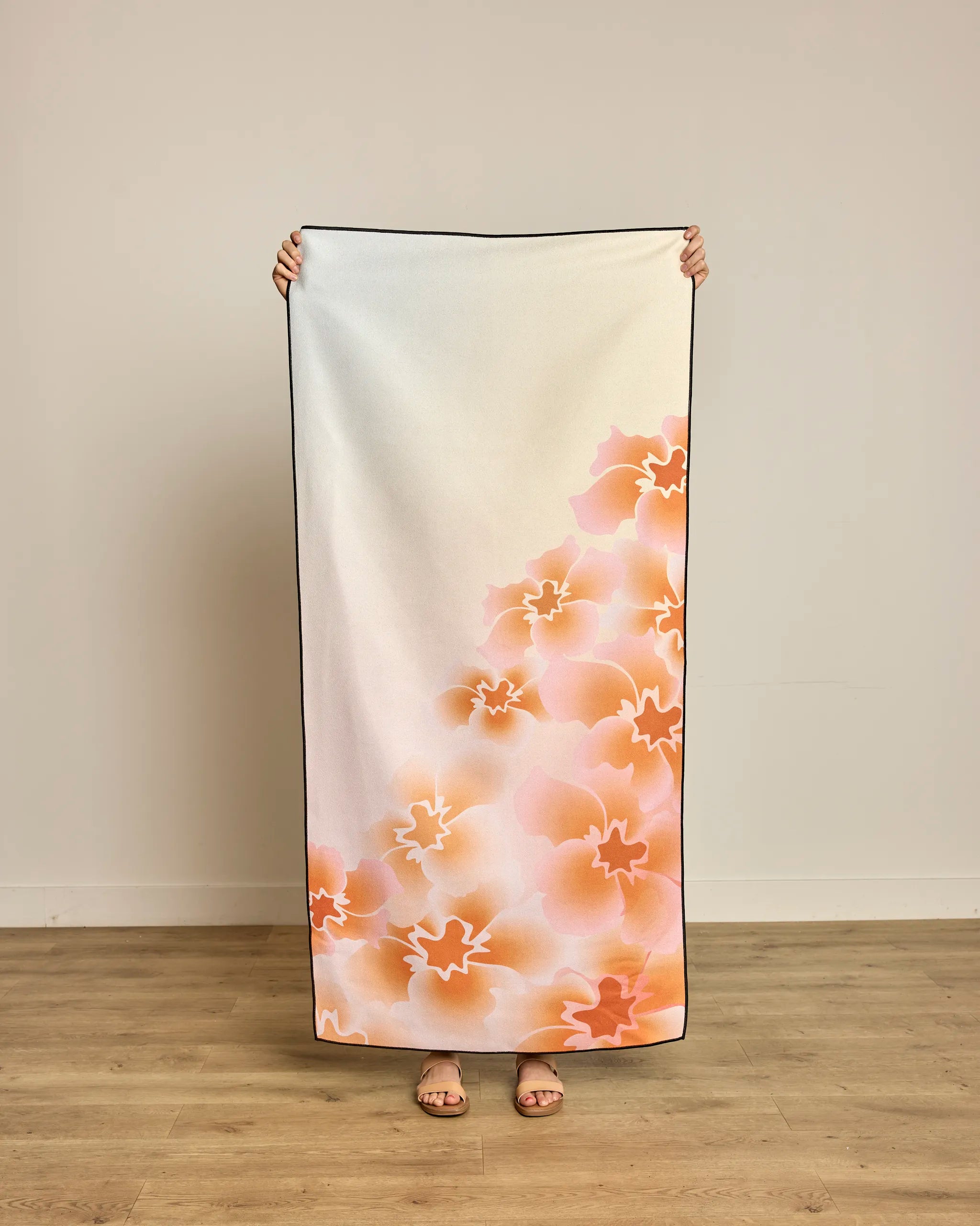 A person stands against a plain wall holding up the Geometry Nicole quick-drying beach towel, featuring an orange and pink floral pattern on a white background; only their hands and feet are visible.
