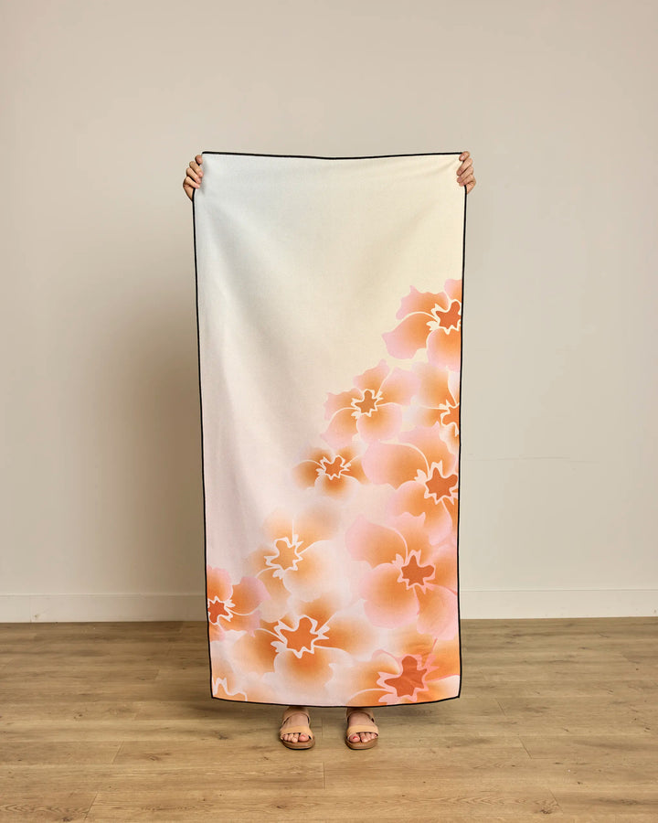 A person stands against a plain wall holding up the Geometry Nicole quick-drying beach towel, featuring an orange and pink floral pattern on a white background; only their hands and feet are visible.