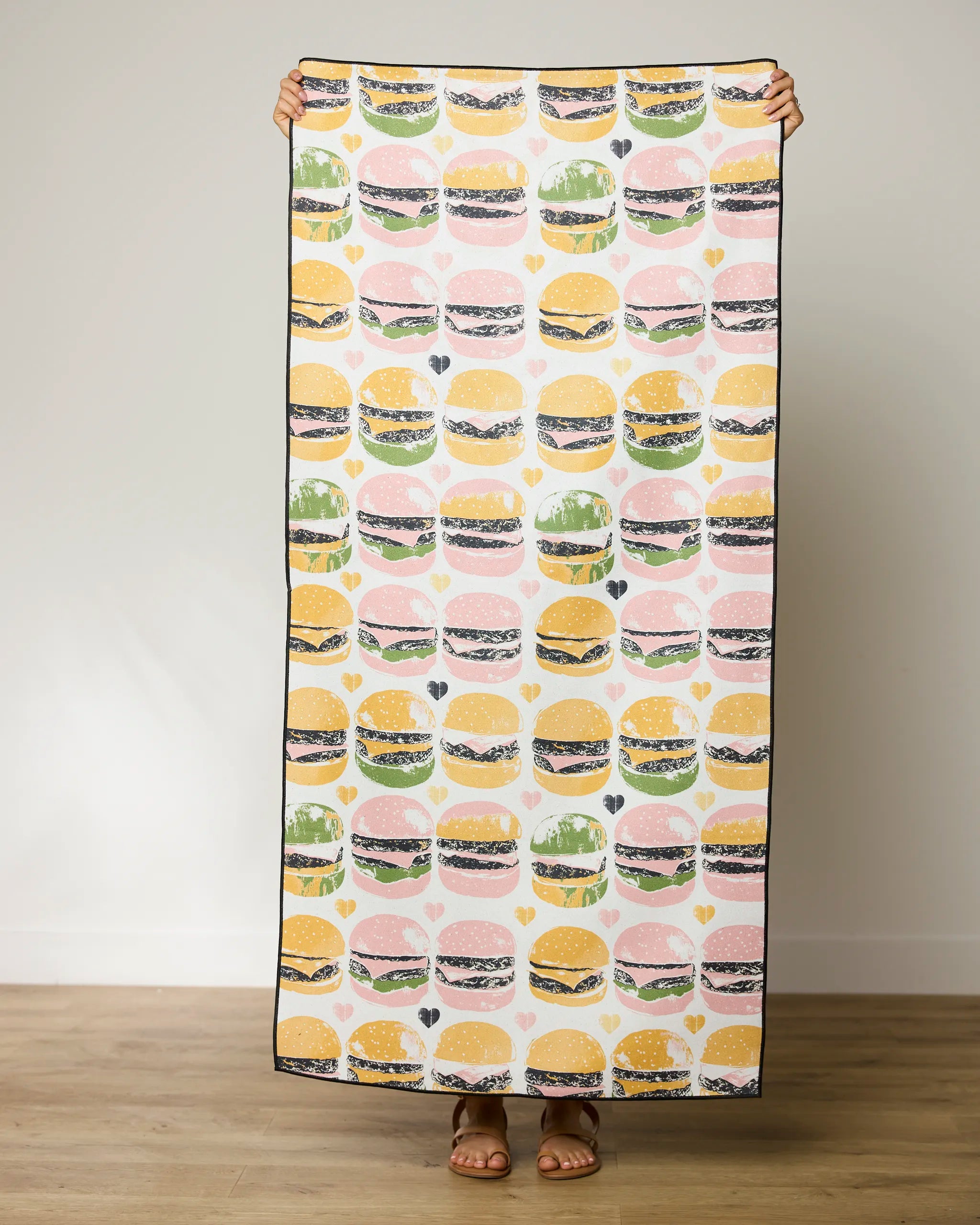 A person holds up a Geometry On The Grill Micro-loop Beach Towel featuring a repeating pattern of colorful hamburgers on a white background, with only their hands and feet visible.