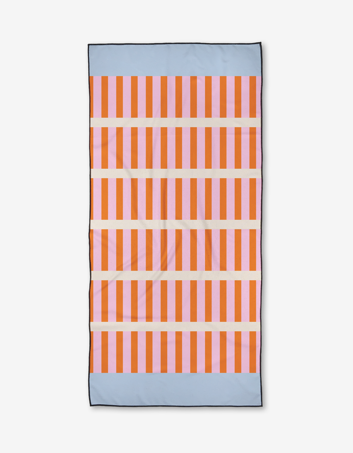 The Red Glow towel by Happy Hemera is a quick-drying beach towel with vertical orange and pink stripes, horizontal blue bands at the top and bottom, and three thin cream stripes crossing the center.