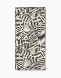The Rhythm Current Stripes towel by Sarah Daugherty is a rectangular beach towel with a black and white geometric pattern of diagonal lines and dots, creating an abstract, repeating design.