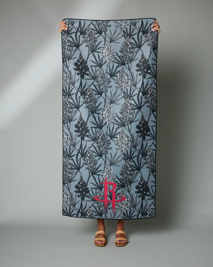 A person holds up the "Rockets Floral" blue recycled beach towel by NBALAB x Geometry, featuring a dark floral pattern and a red monogram near the bottom. Only their hands and feet in sandals are visible against a plain gray background.