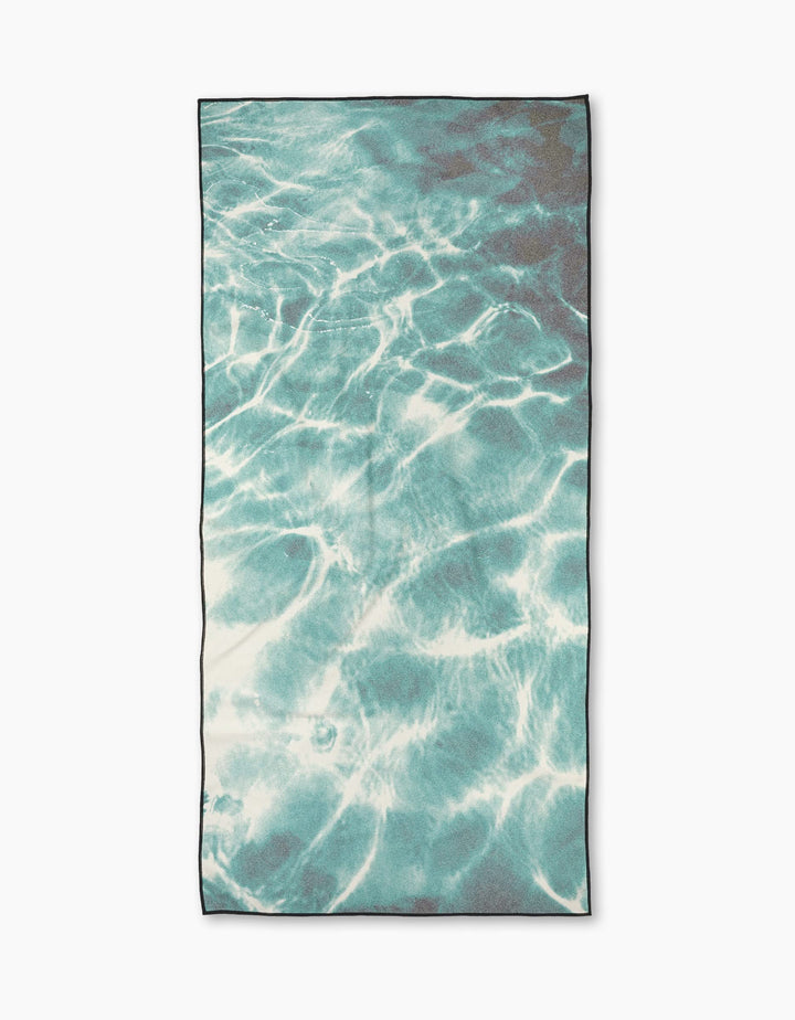 The Geometry Saltwater Glow towel is a rectangular, quick-drying towel featuring a printed design of blue water with ripples and sunlight reflections, mimicking a swimming pool surface.