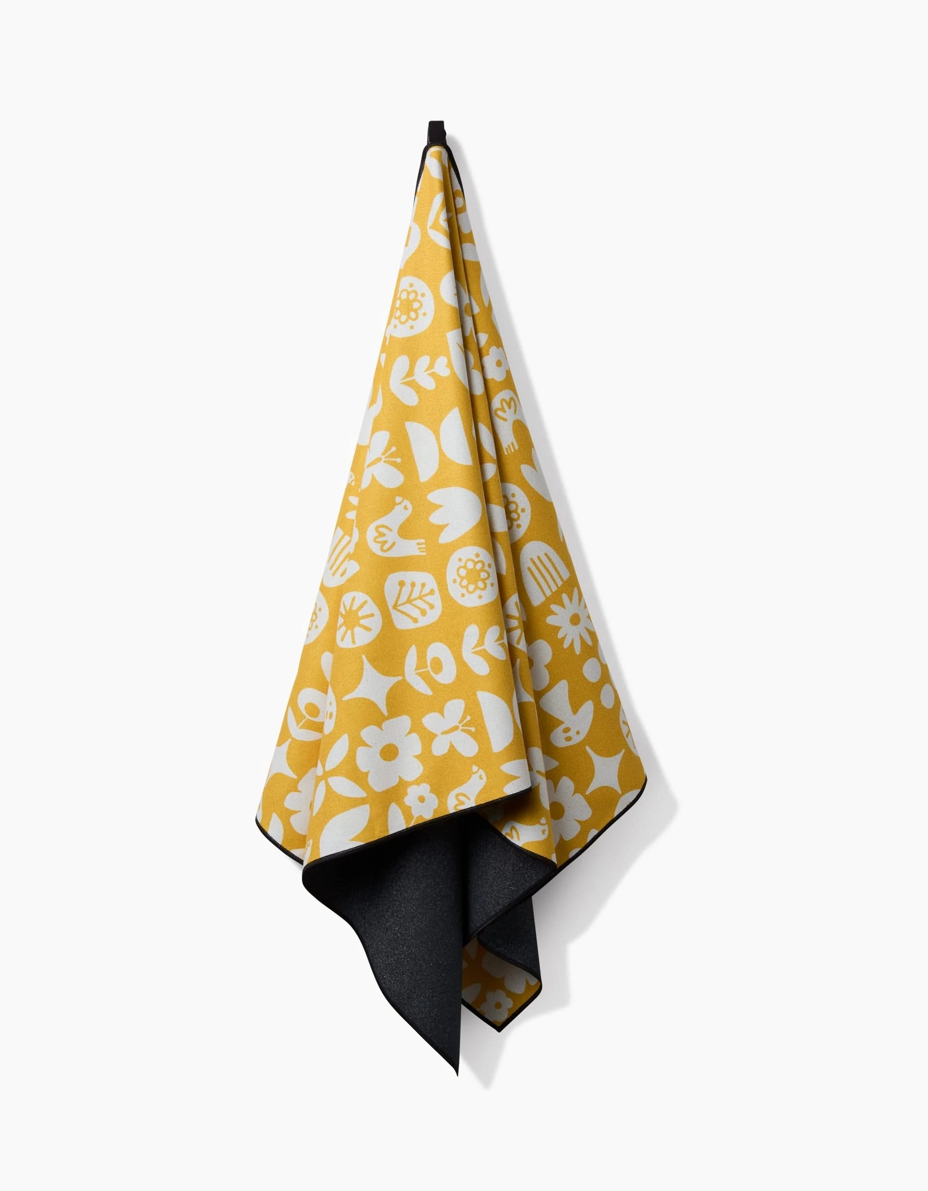 The Kayla Ann Scandi Spring beach towel, featuring yellow with white floral and botanical patterns and a dark gray underside, hangs from a black hook against a plain white background. It’s crafted from quick-drying materials.