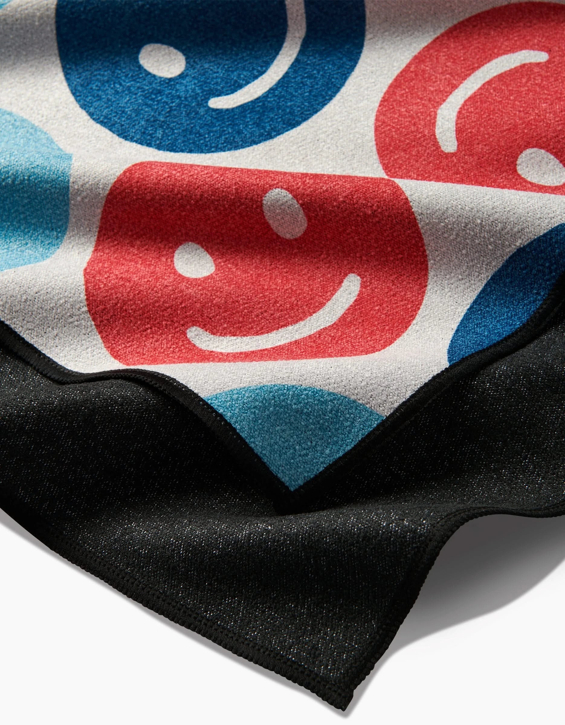 A close-up of the Smiley Face July beach towel by Hufton Studio, made from recycled materials, shows red and blue happy and sad faces on white with a solid black border.