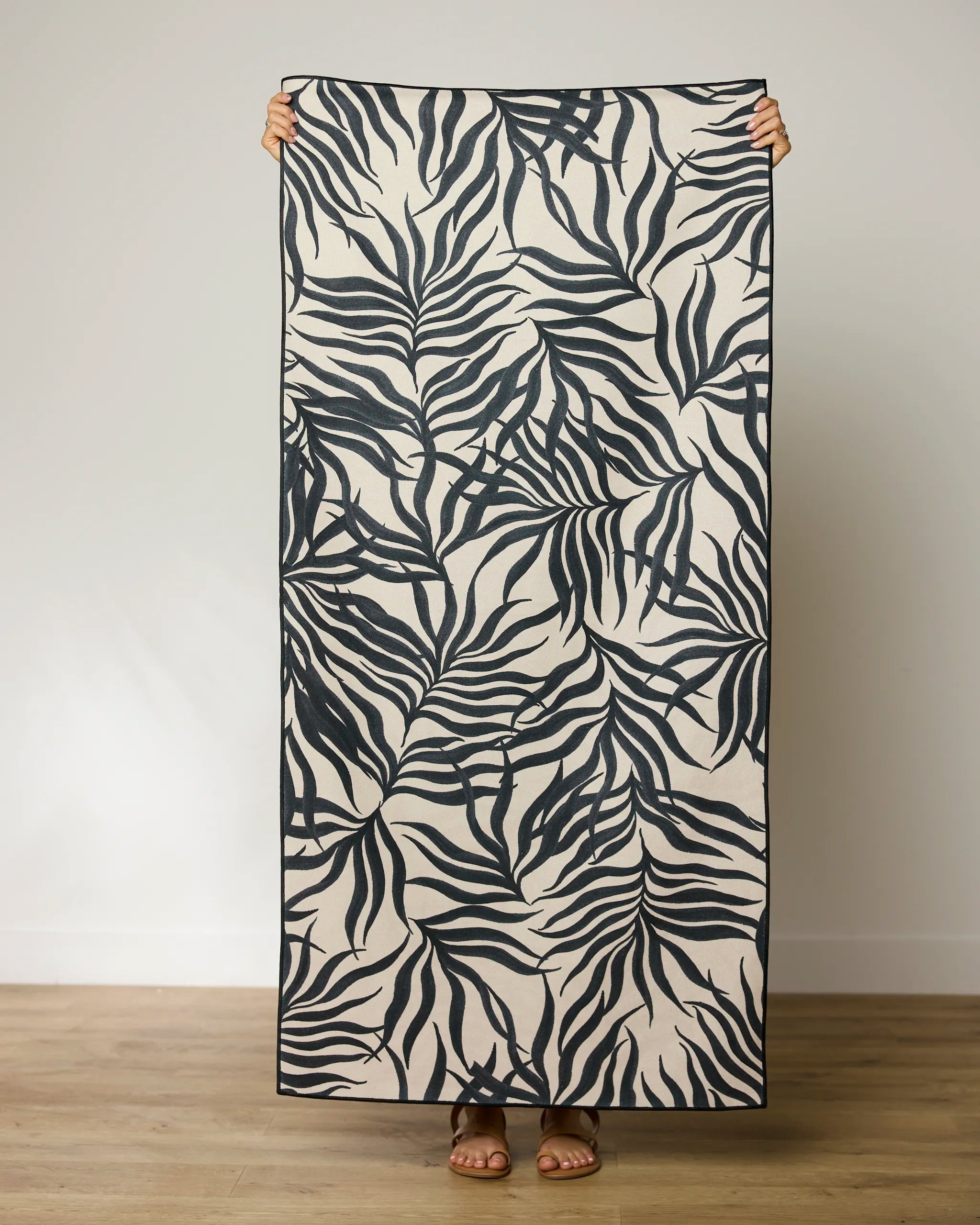 A person holds up Geometry’s "Solar Flare" beach towel—a quick-drying, rectangular towel with a bold black and white abstract leaf pattern—while standing on a light wooden floor against a plain white wall, showing only hands, feet, and sandals.