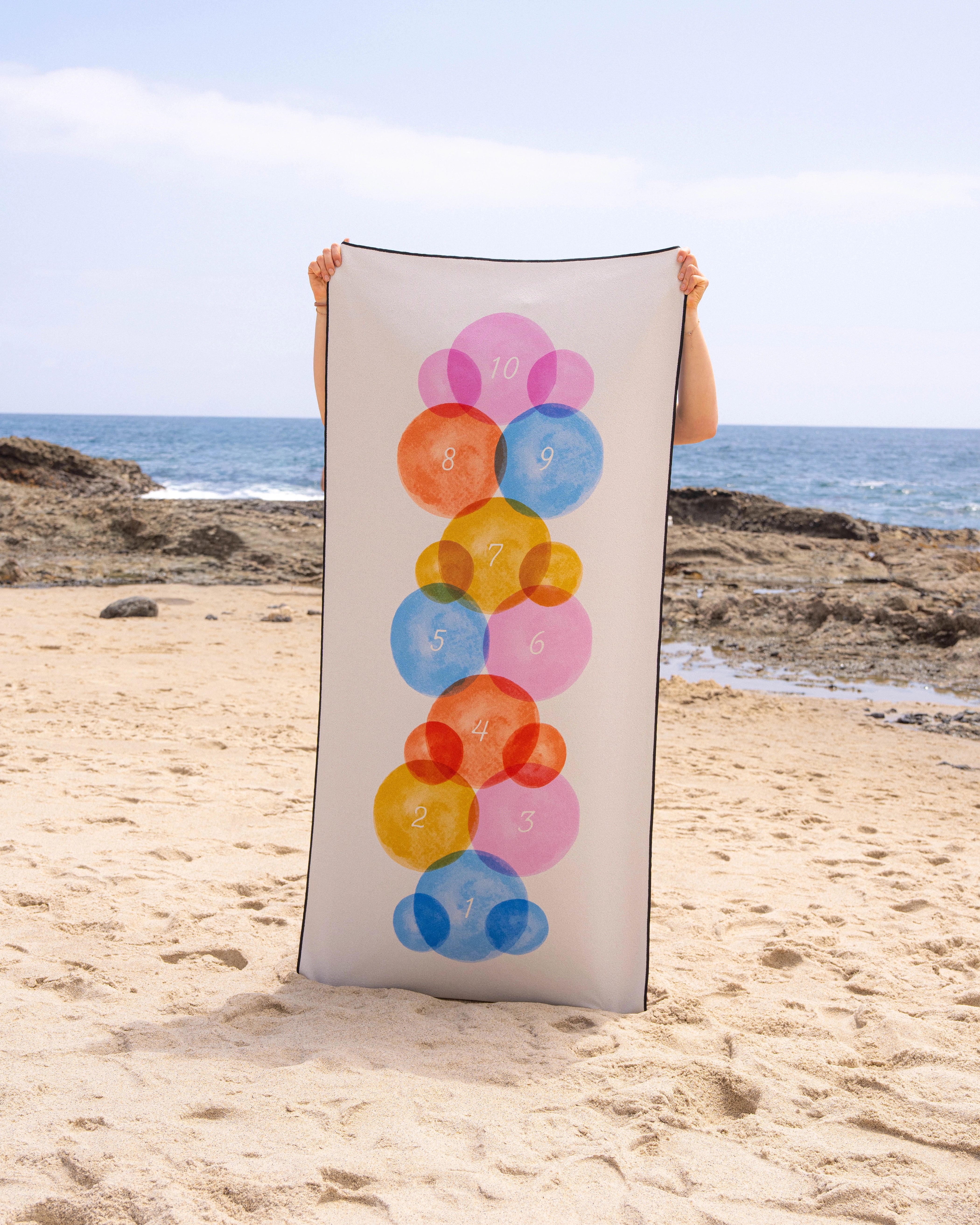 A person stands on a sandy beach holding Geometry’s Stepping Stones quick-drying micro-loop towel, featuring colorful overlapping circles numbered 1–10, with the ocean and rocky shoreline in the background.