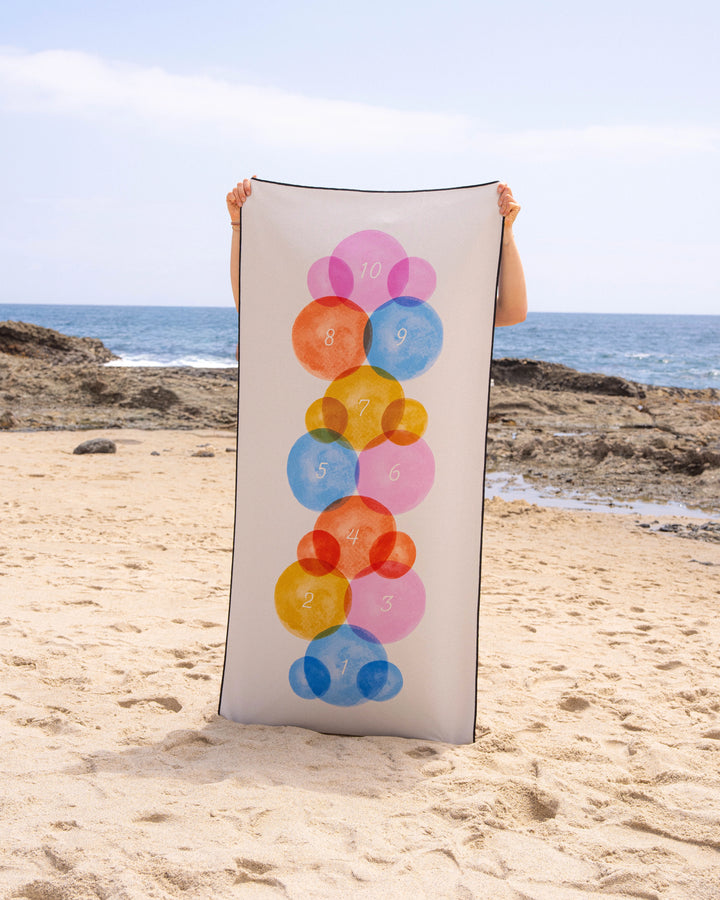 A person stands on a sandy beach holding Geometry’s Stepping Stones quick-drying micro-loop towel, featuring colorful overlapping circles numbered 1–10, with the ocean and rocky shoreline in the background.