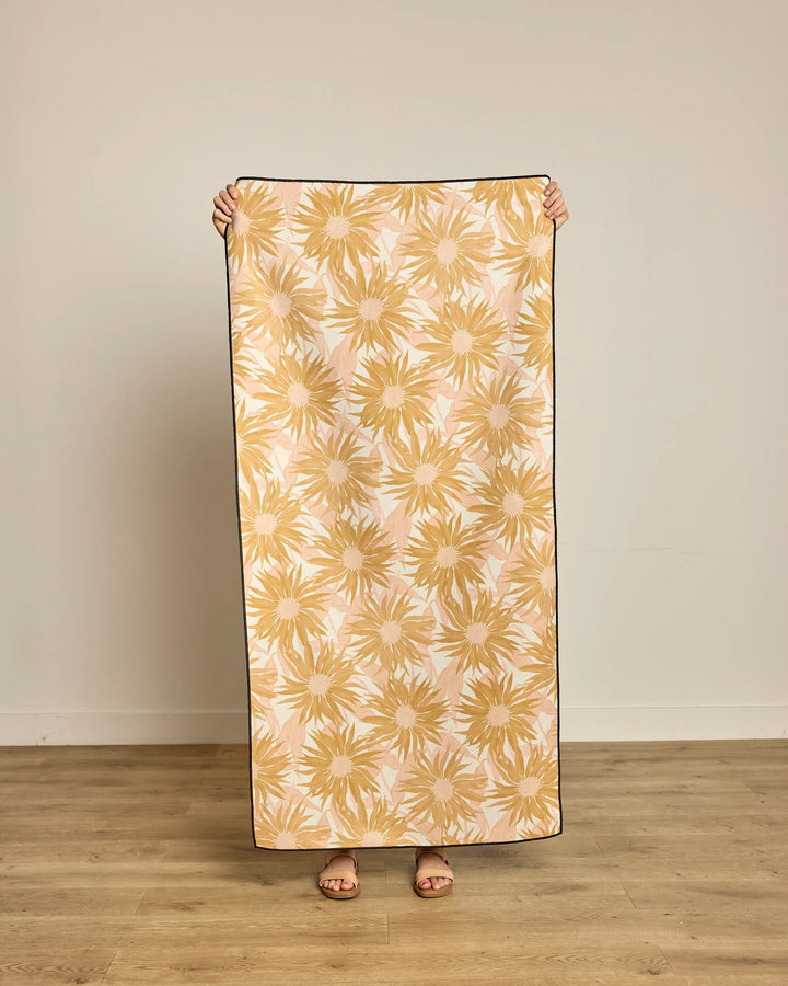 A person indoors on a wooden floor holds up the Geometry Sundara Micro-loop Beach Towel, featuring a yellow and white floral pattern that covers their face and upper body.