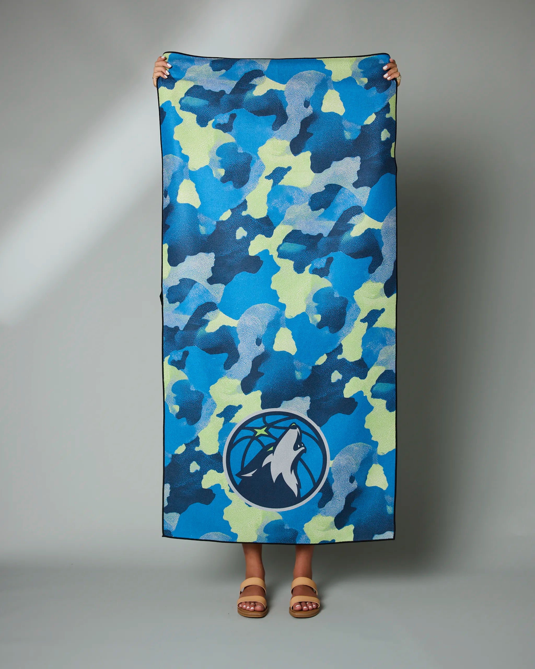 A person displays the Timberwolves Camo towel by Geometry, featuring blue and green camouflage and a Minnesota Timberwolves logo at the bottom; only their hands and feet are visible.