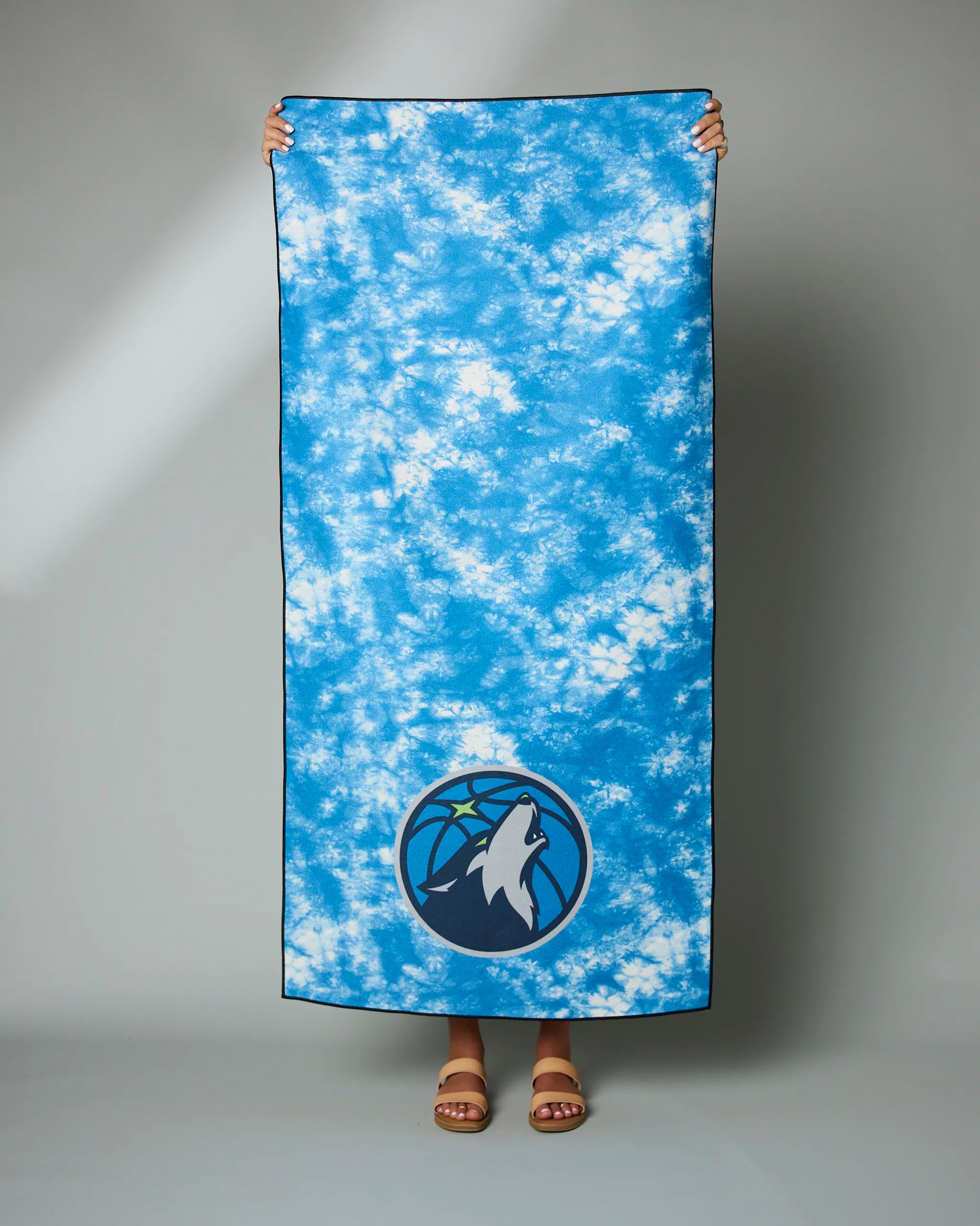 A person holds up the Geometry Timberwolves Flash towel, a large blue tie-dye design with the Minnesota Timberwolves logo near the bottom; only their hands and sandal-clad feet are visible.